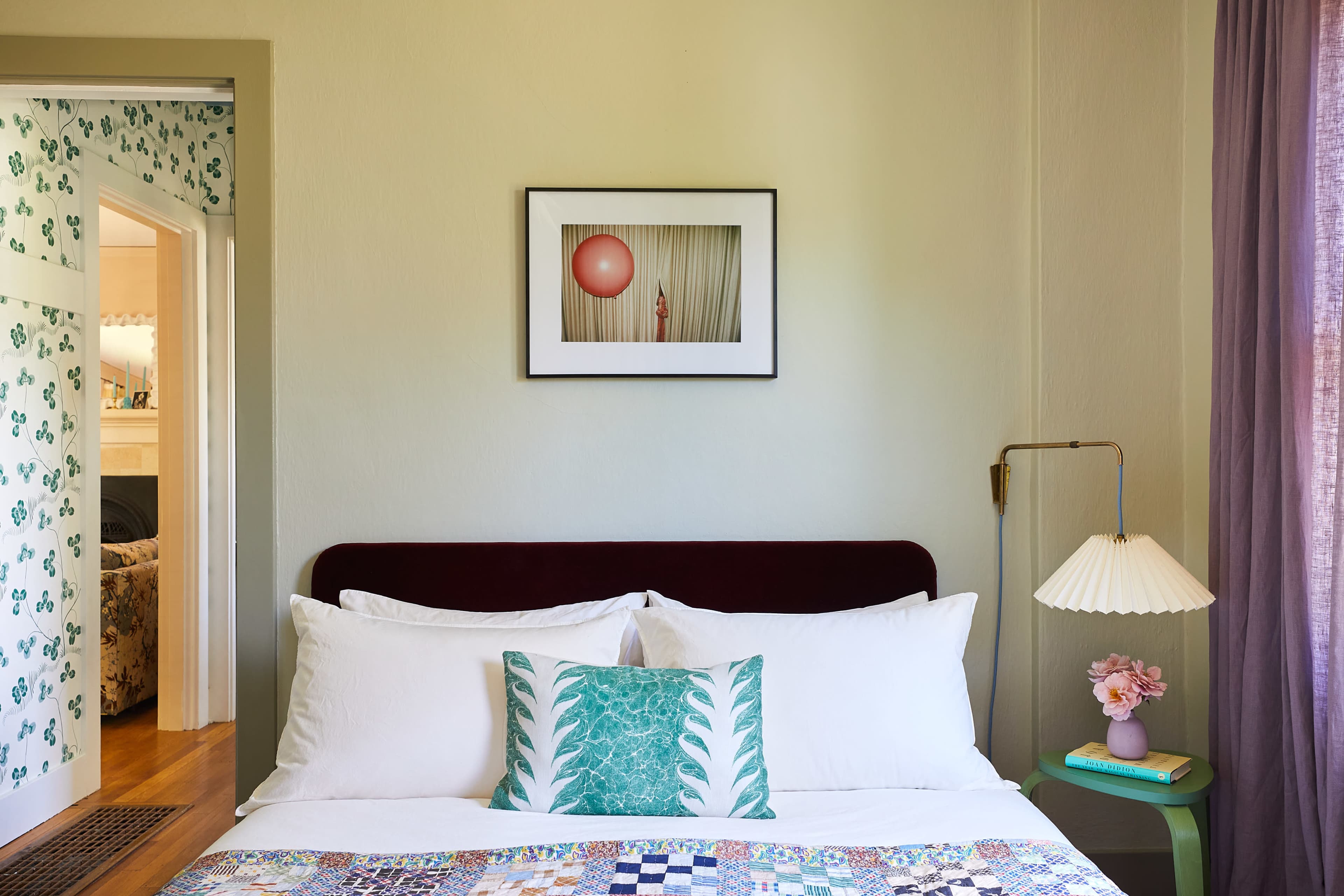 A neatly arranged bed with white linens and a decorative pillow is placed against a pale green wall, featuring a framed artwork and a bedside lamp.
