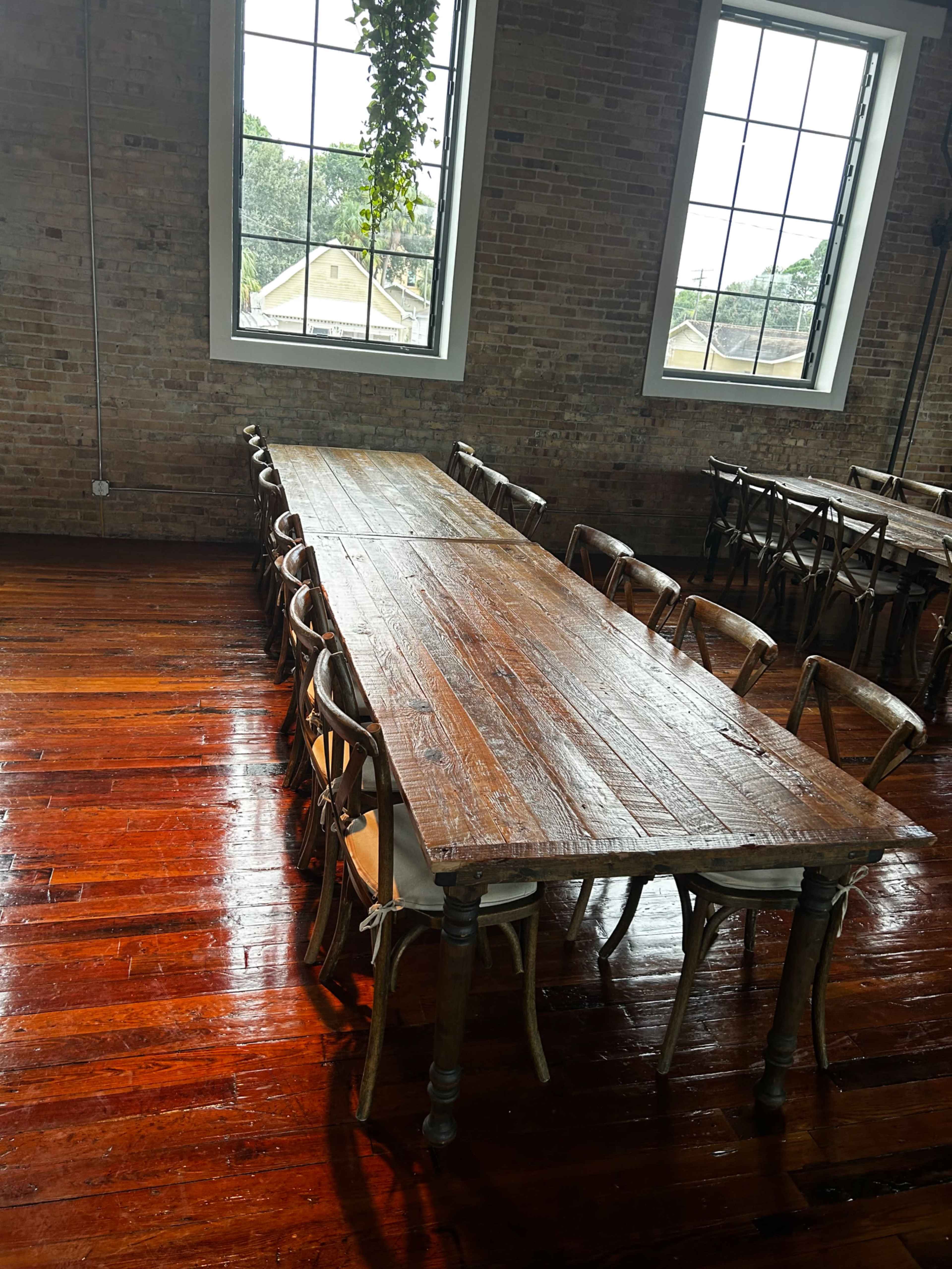 A long wooden table with several chairs is positioned in a room with large windows and hardwood floors.