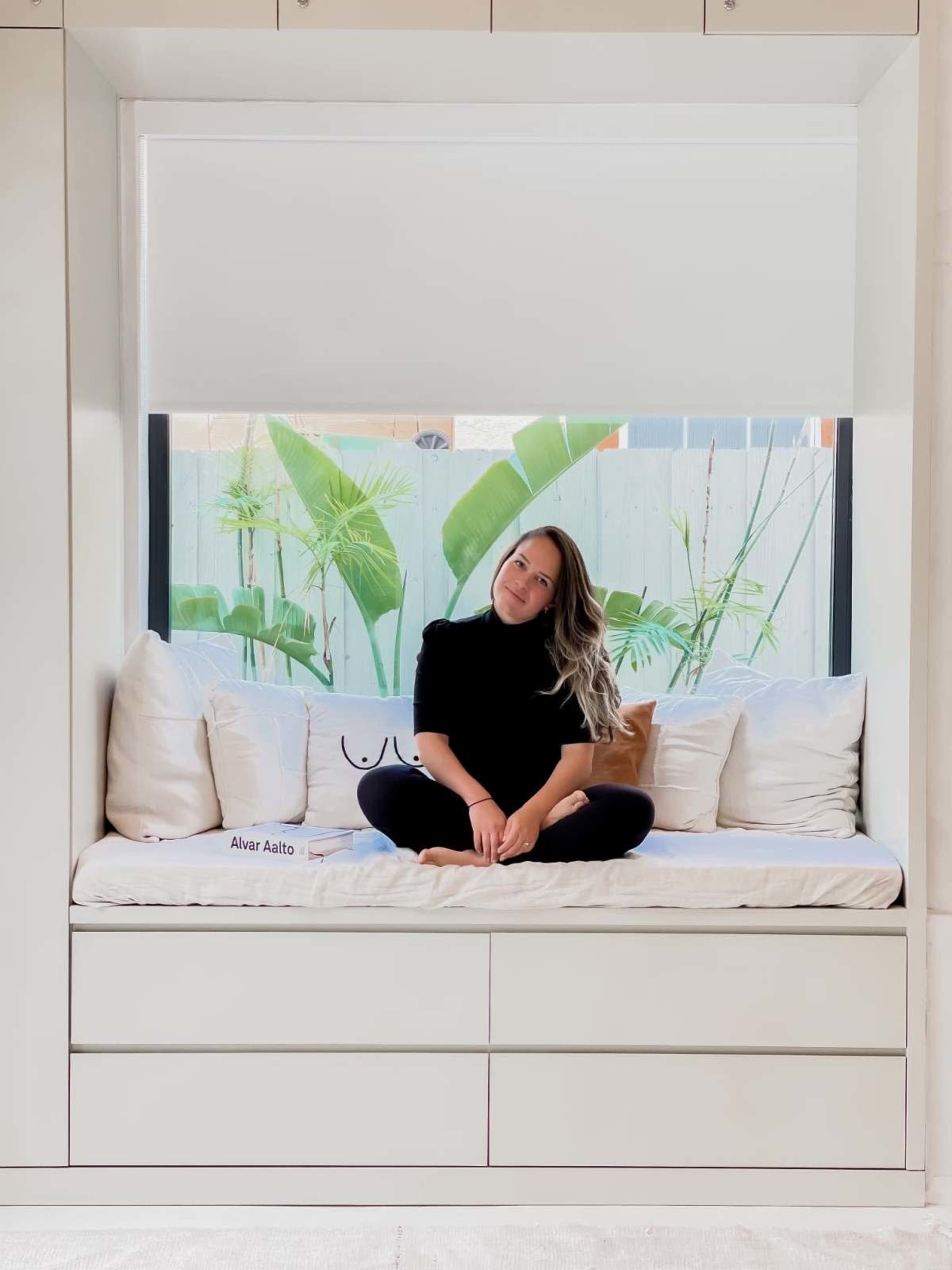 A person sits cross-legged on a window seat surrounded by potted plants and decorative pillows.
