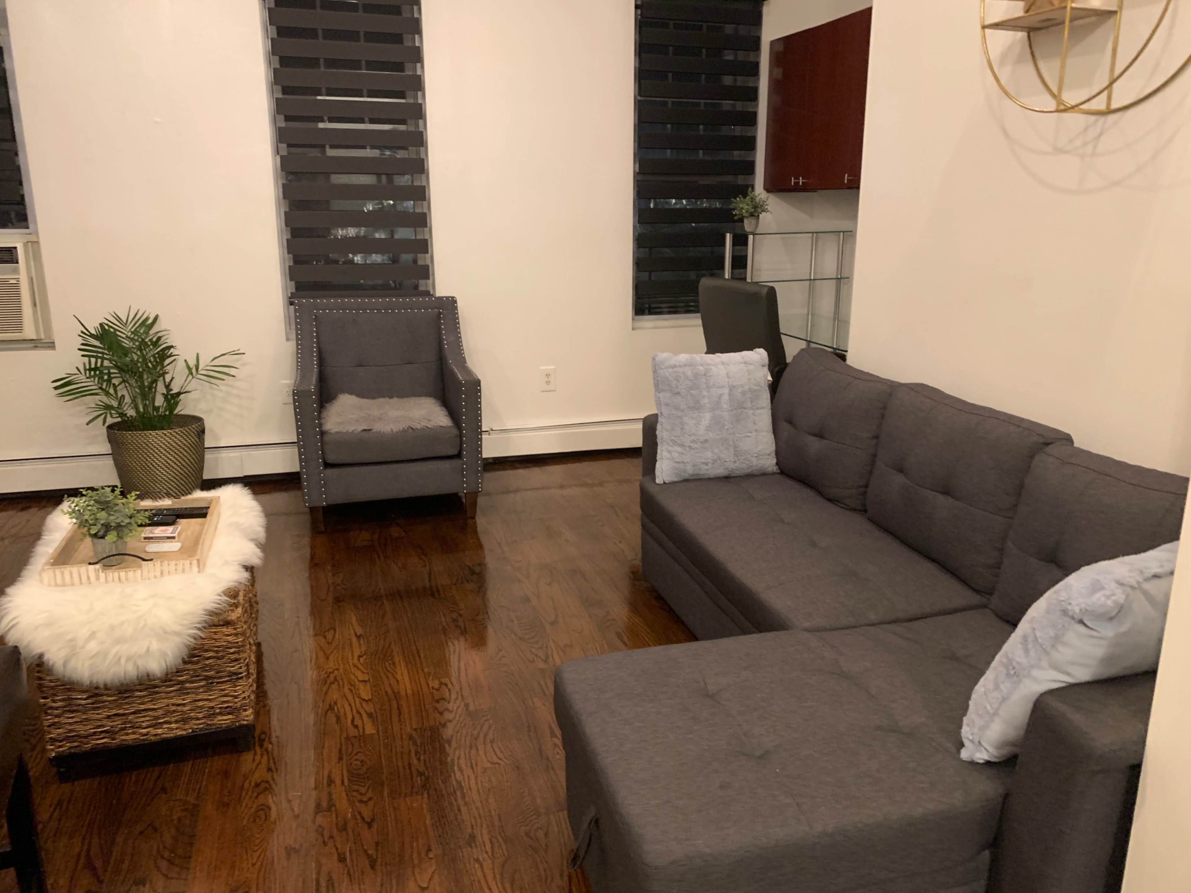 A modern living room with a gray sectional sofa, a textured coffee table, and a single armchair, all set against a backdrop of dark window shades.