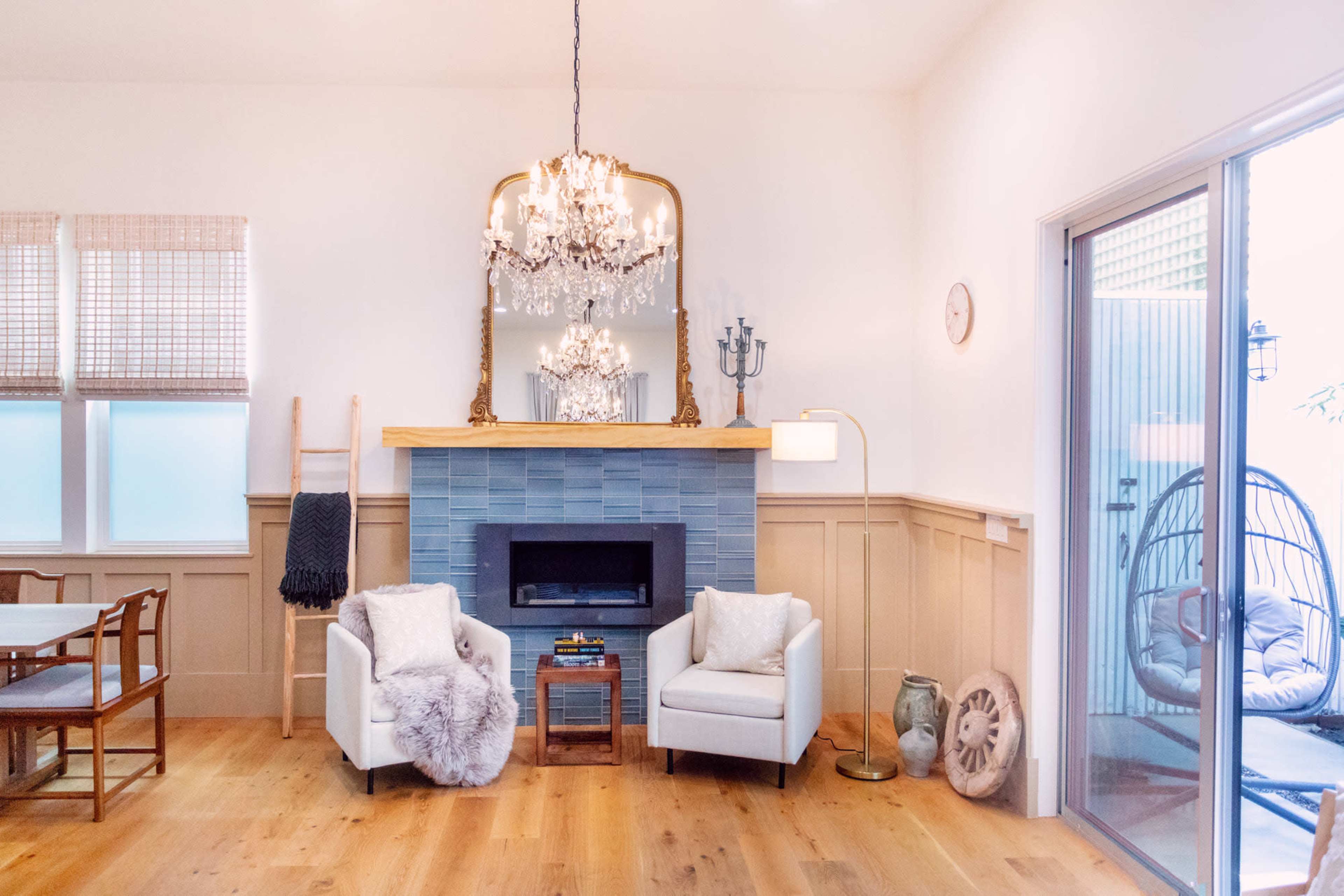 A bright living room features two white armchairs with plush cushions, a blue tiled fireplace, a large mirror, and a hanging chair.