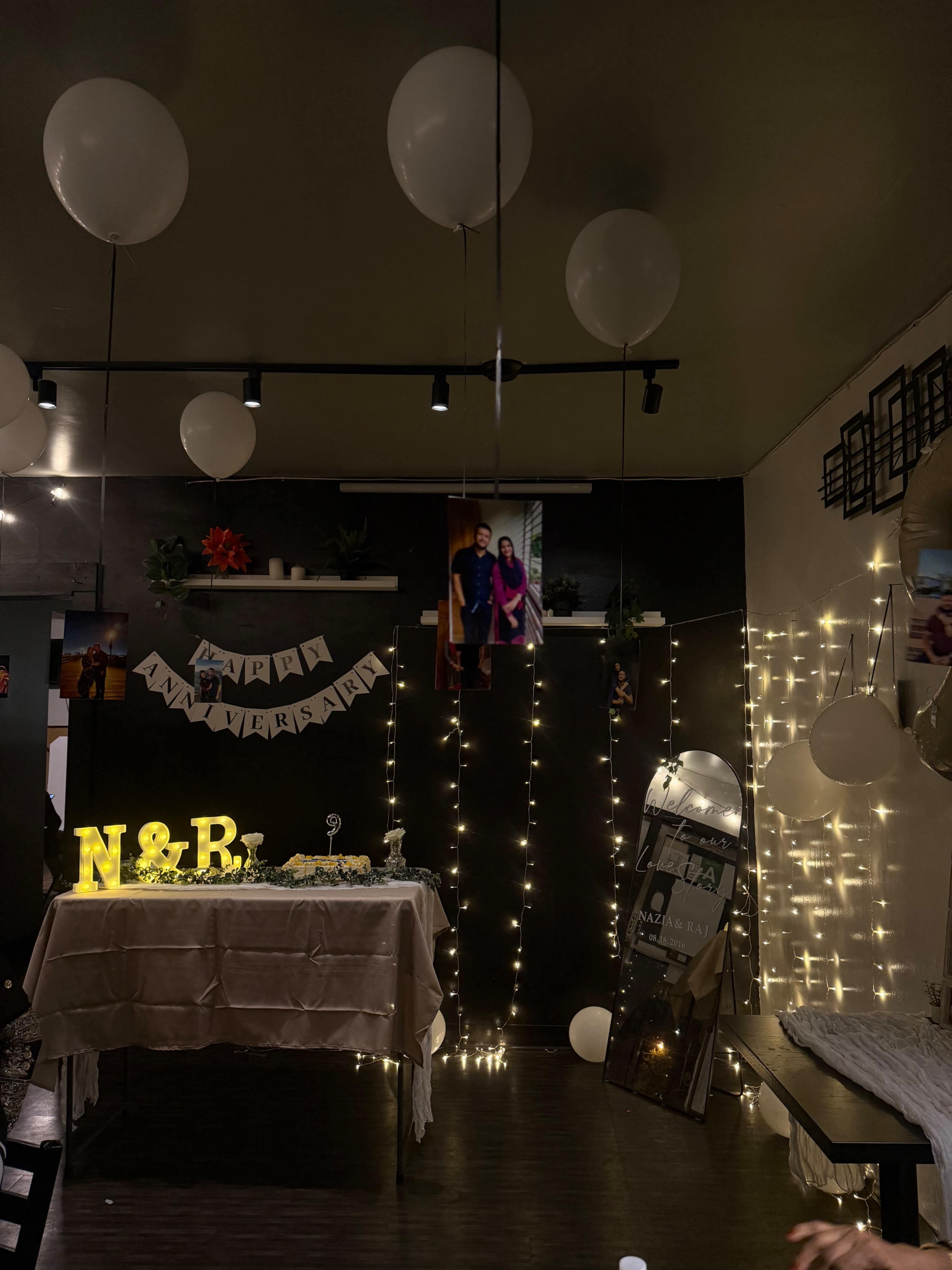 The image shows a decorated room with a table in the center, string lights, balloons, and a banner, creating a festive atmosphere.