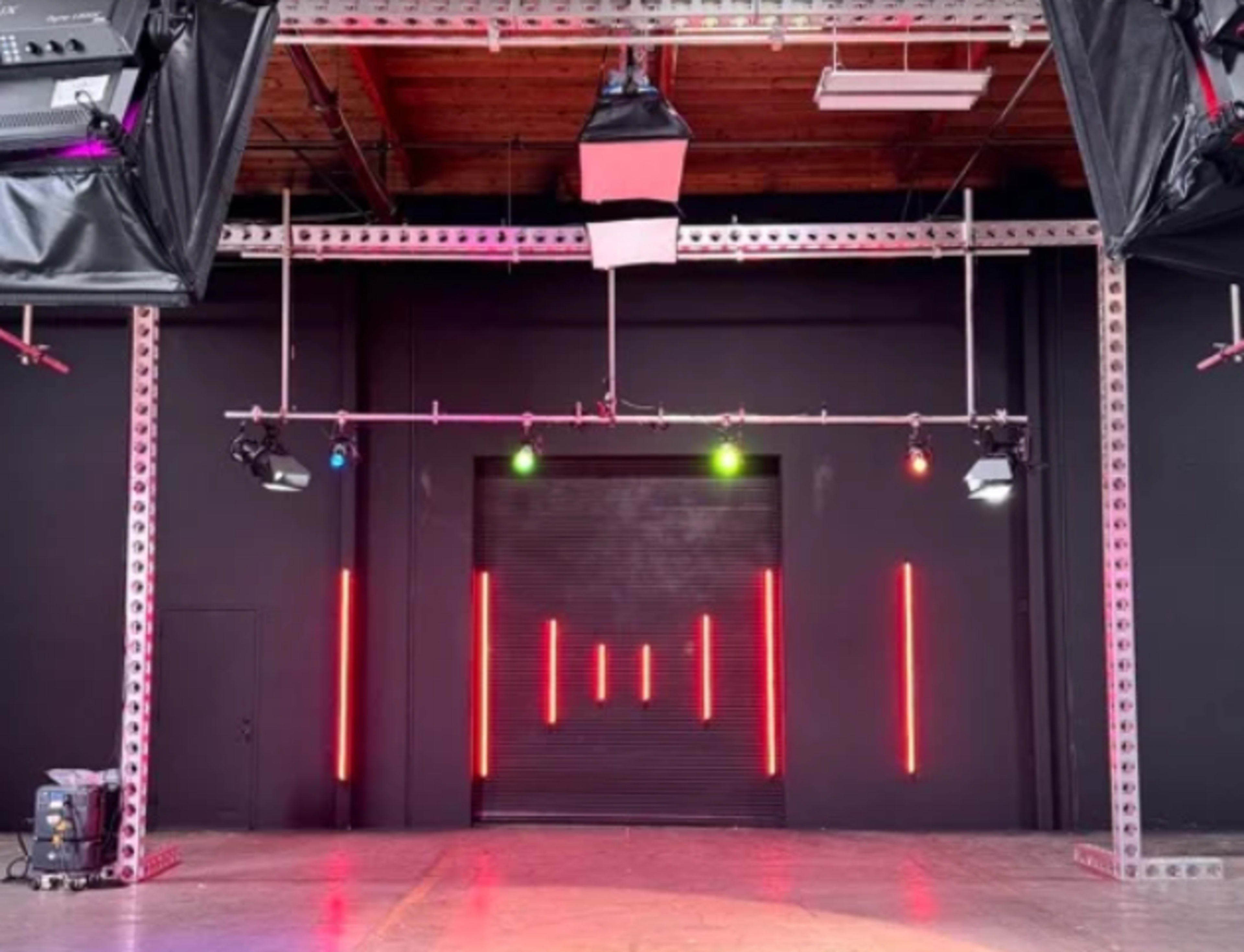 The image shows a studio space with a black backdrop, featuring overhead lighting rigs and vertical LED strips illuminating the walls.