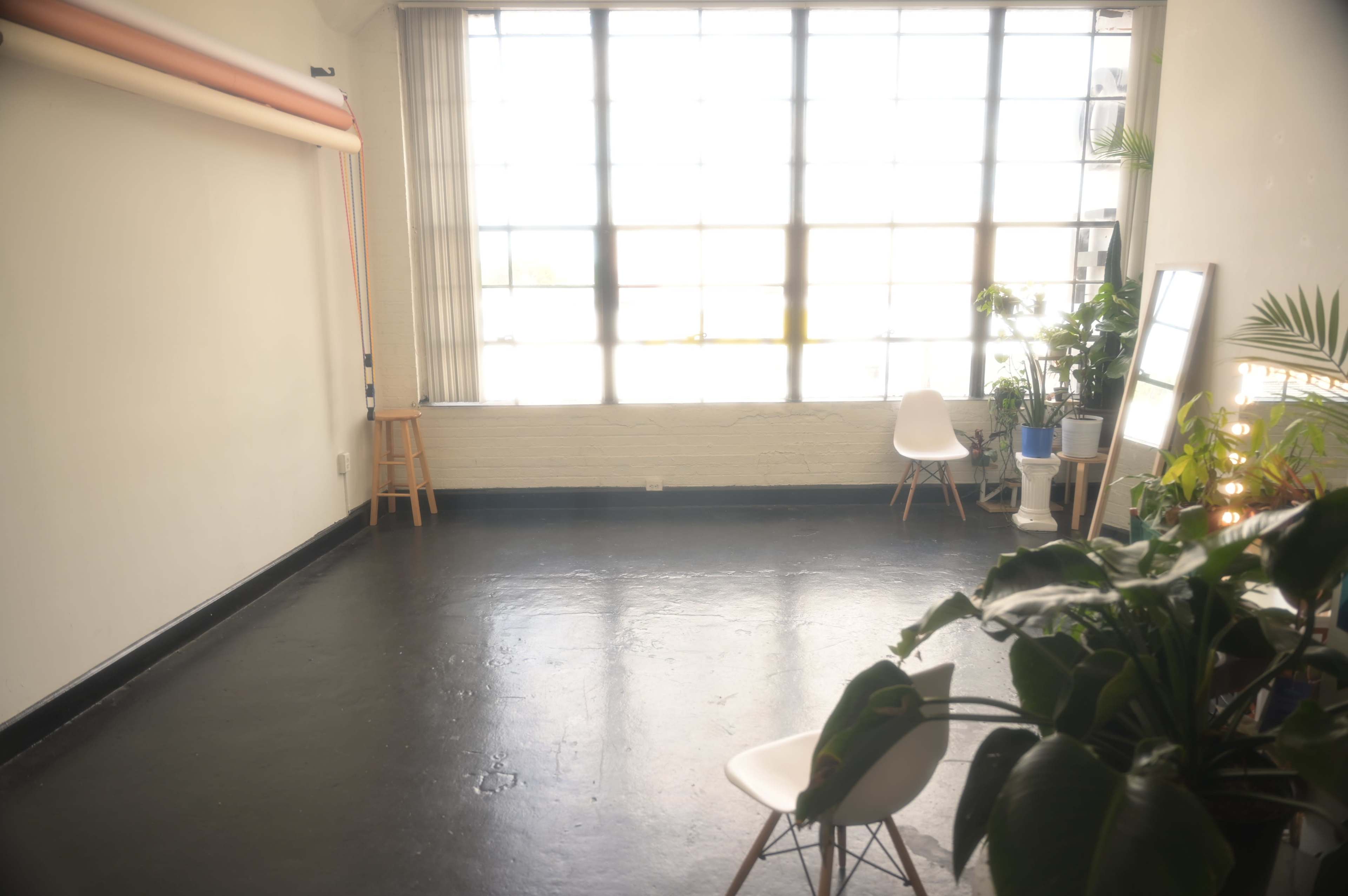 A spacious room with a large window, a stool, a chair, a mirror, and several indoor plants.