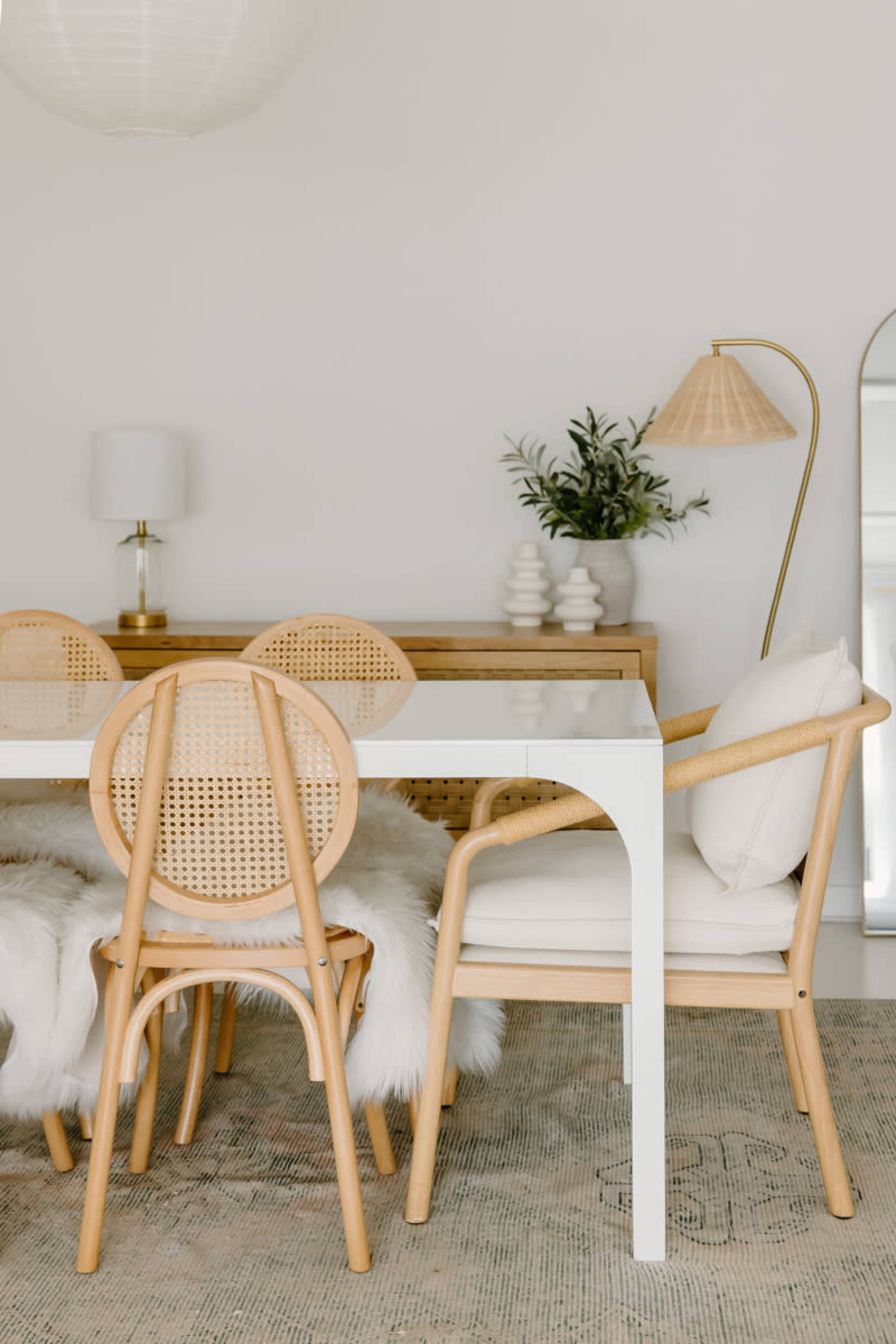 A dining area features a white table surrounded by rattan and wooden chairs, with a sideboard, a lamp, and decorative elements in a neutral color palette.
