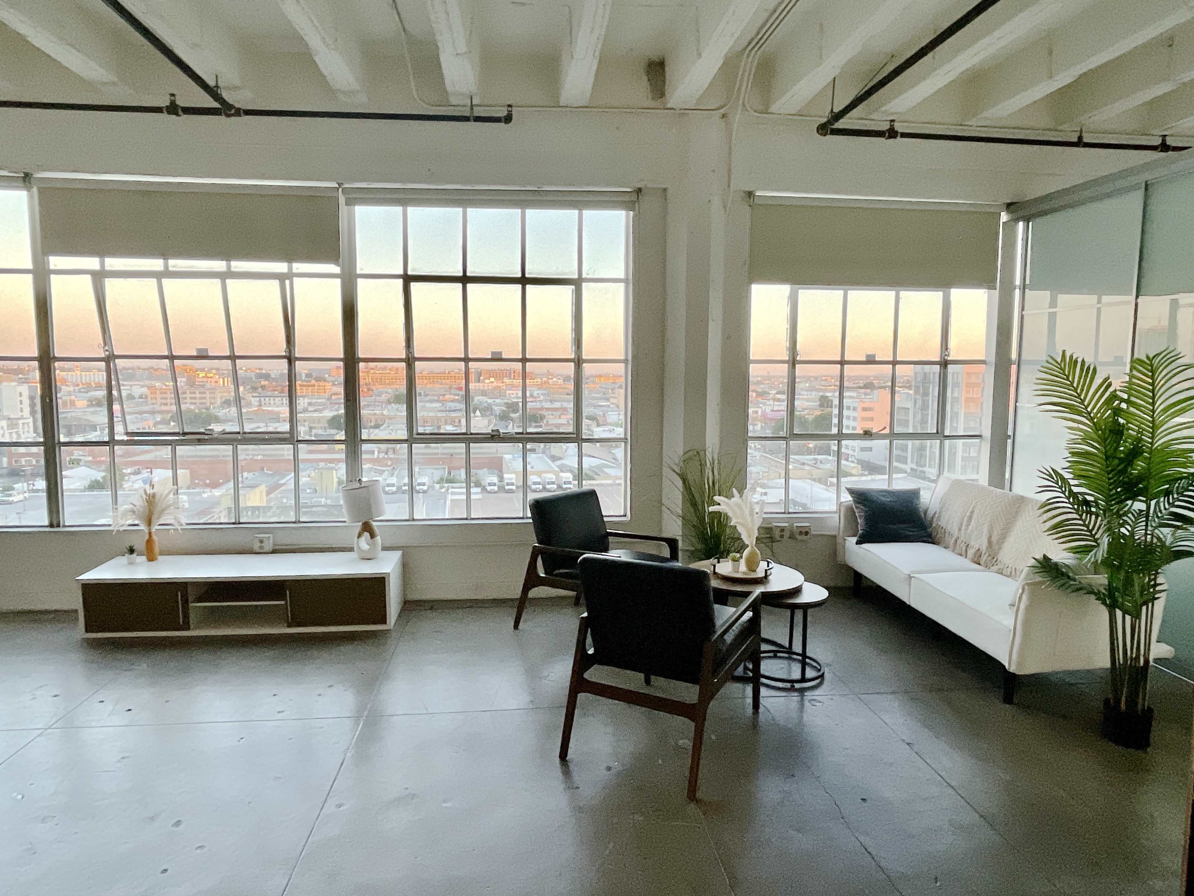 A modern, airy interior with large windows providing a view of a cityscape at sunset, featuring minimalist furniture including a sofa, chairs, and a coffee table.