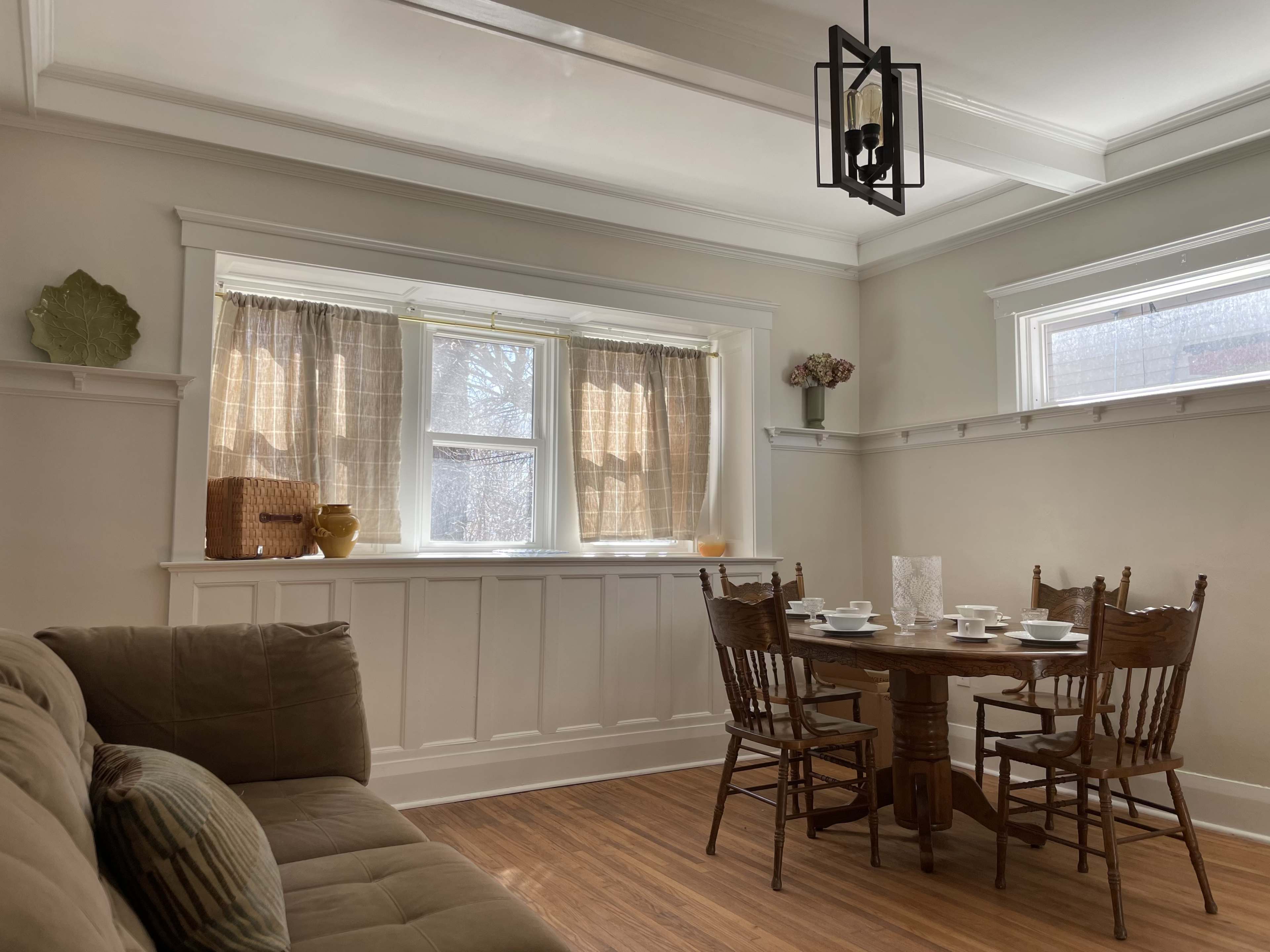 A dining area features a round wooden table set for four, surrounded by chairs, with a couch and window curtains in the background.