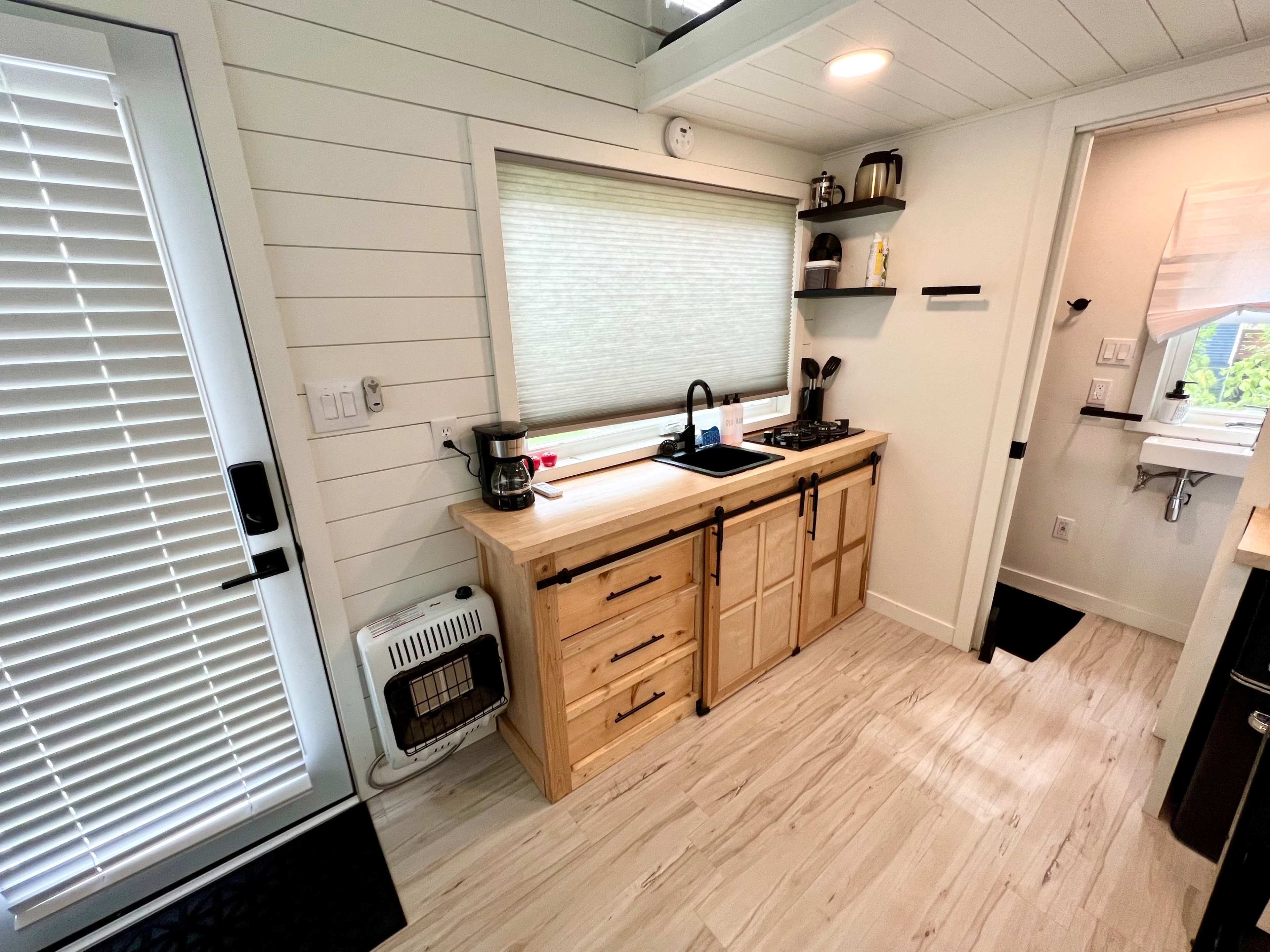 A compact kitchen features wooden cabinetry, a sink, a stovetop, and a window with blinds, all within a well-lit space.