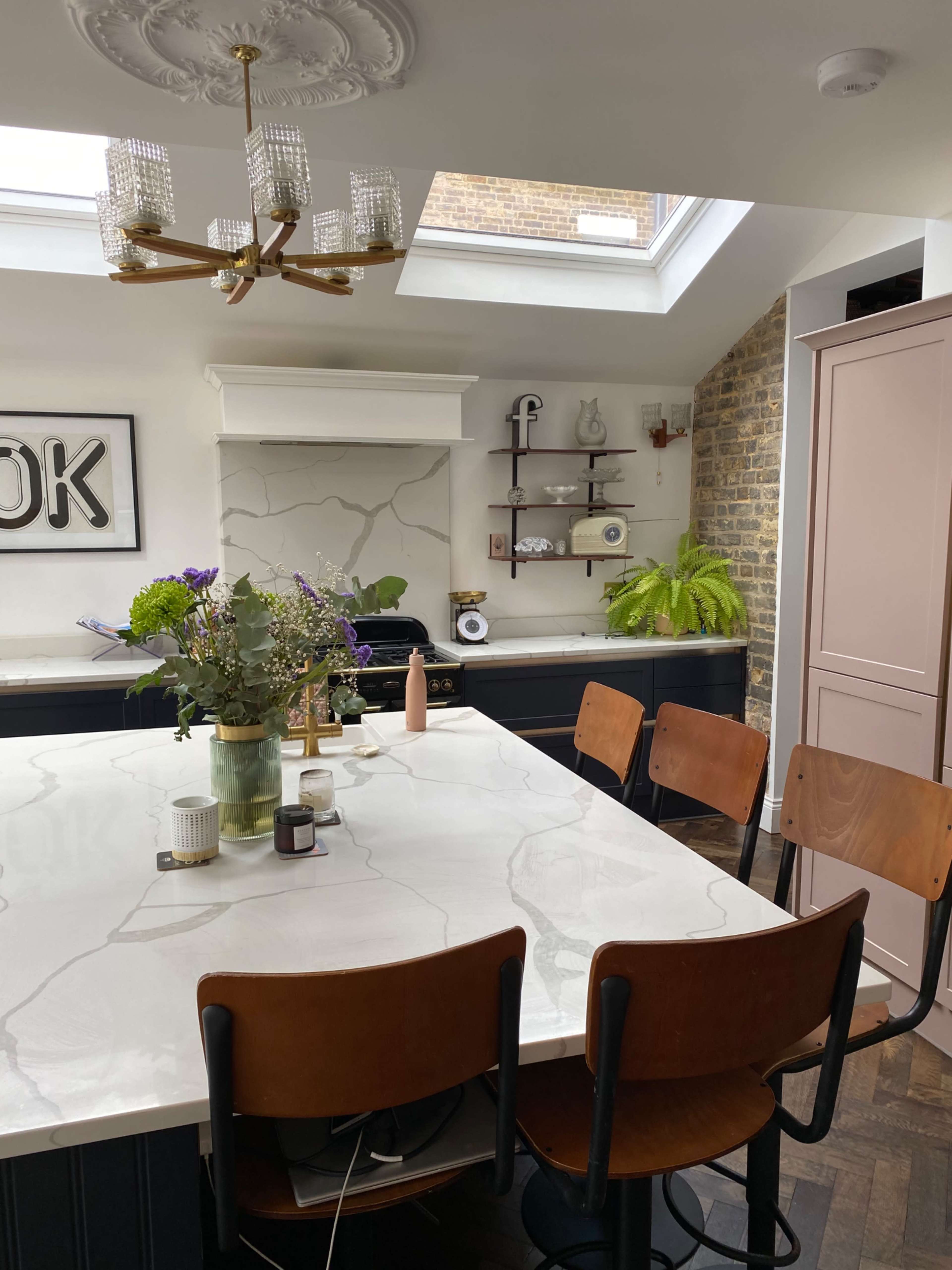 The image shows a modern kitchen with a large marble island, wooden chairs, and a skylight providing natural light.