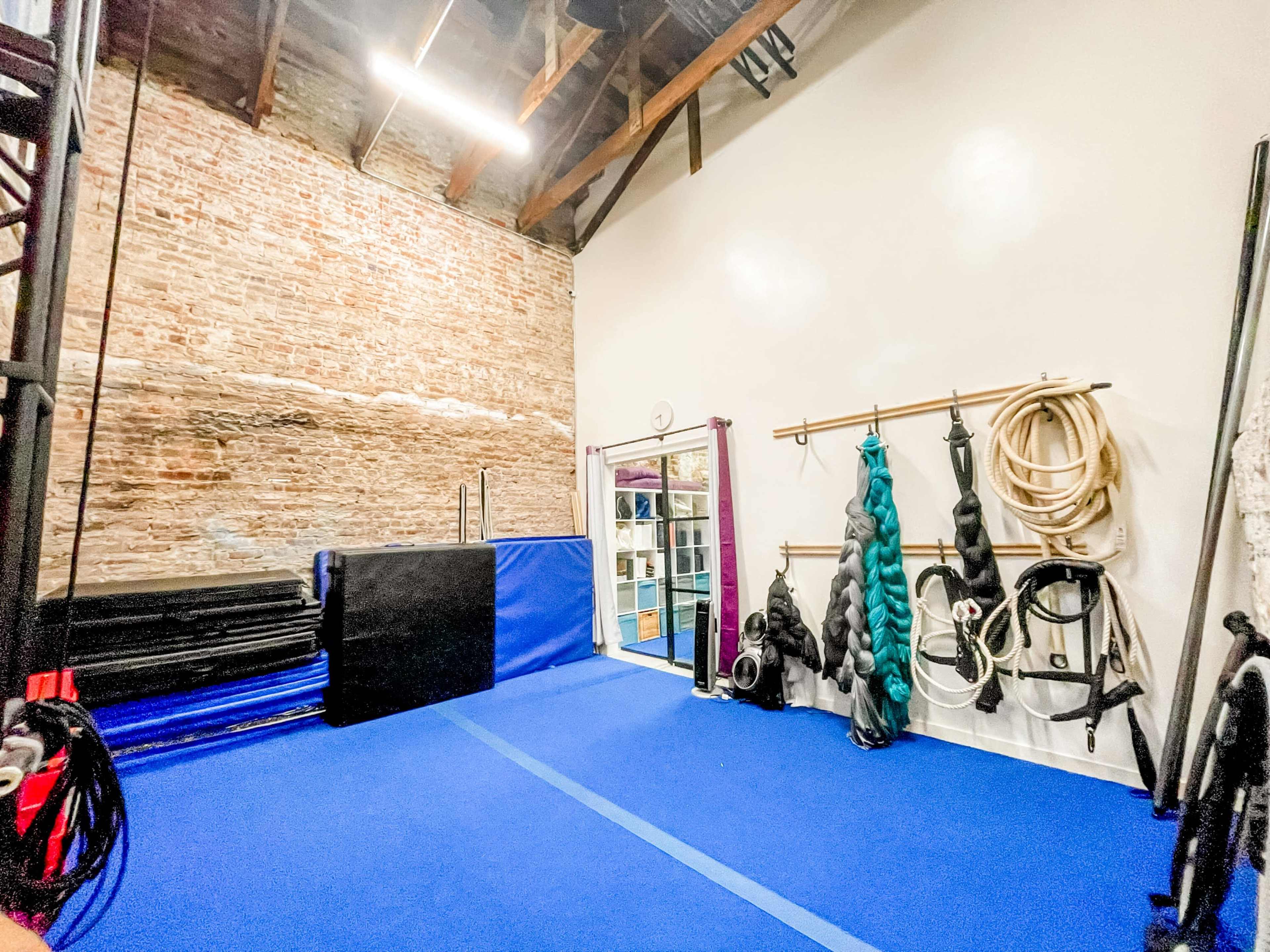 A training space featuring blue flooring, gym mats against a brick wall, and various equipment hanging on a rack.