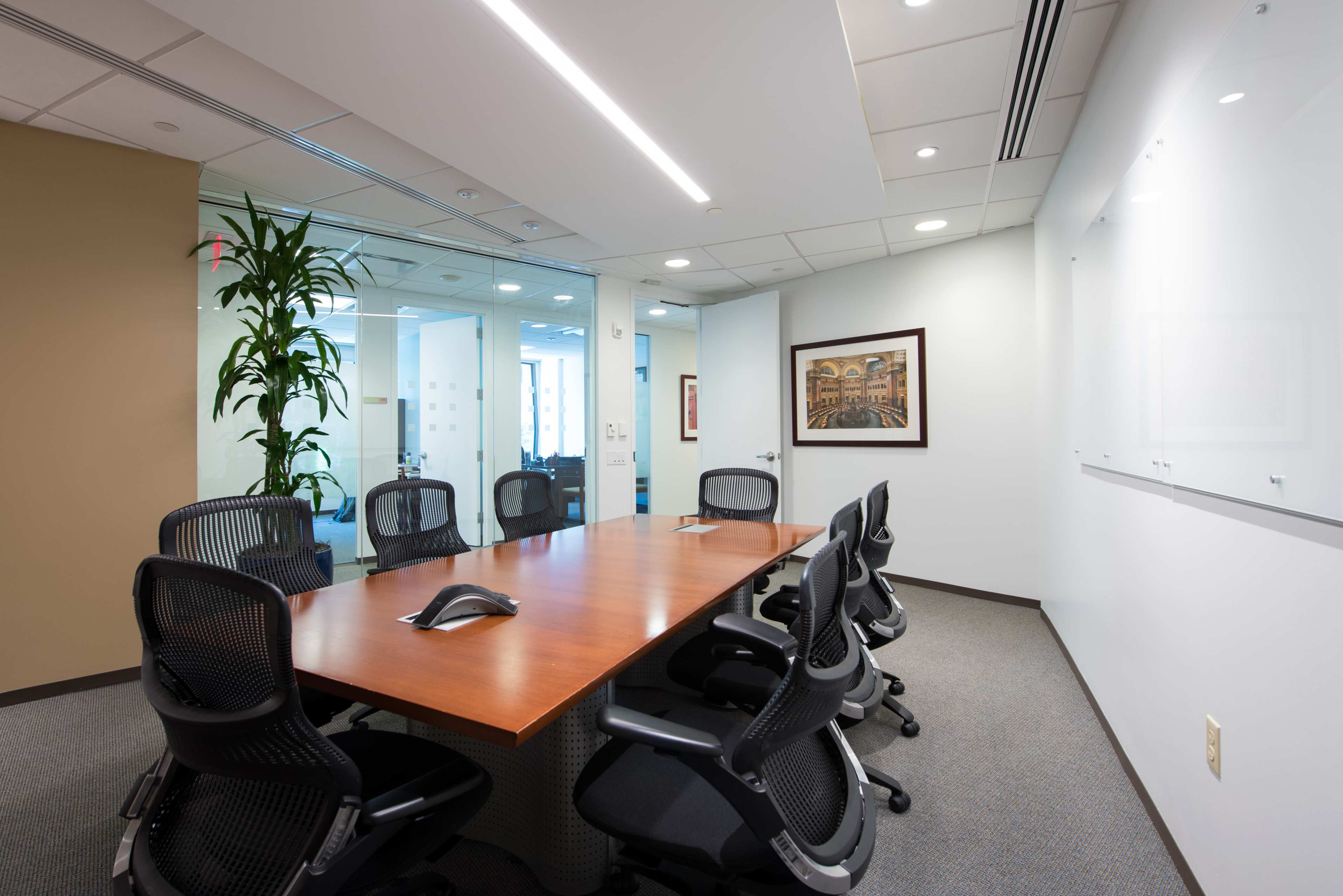 A modern conference room with a large wooden table surrounded by black ergonomic chairs and a glass wall in the background.