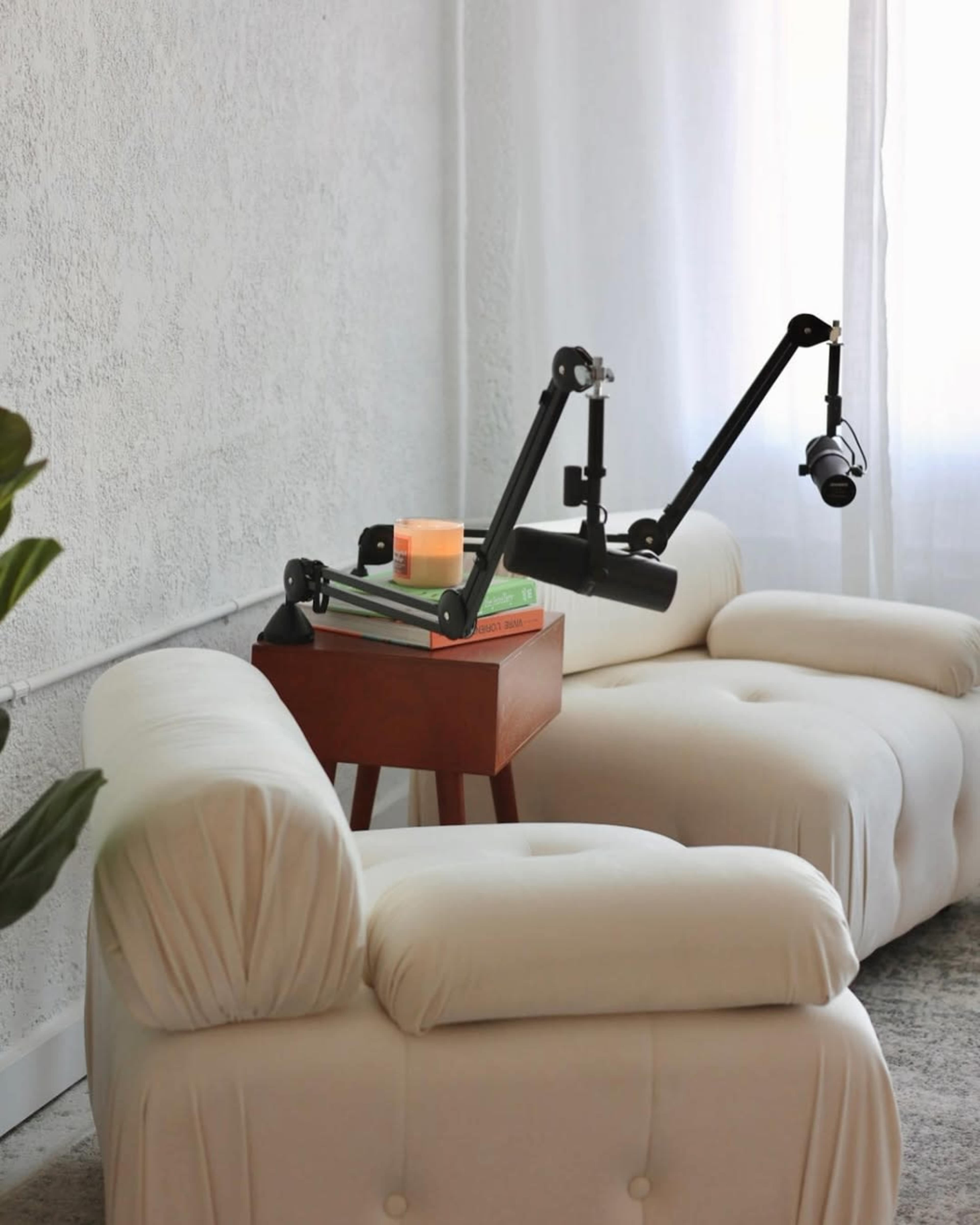 A light-colored, tufted sofa is positioned next to a small wooden side table with stacked books and a candle, illuminated by natural light from a nearby window.