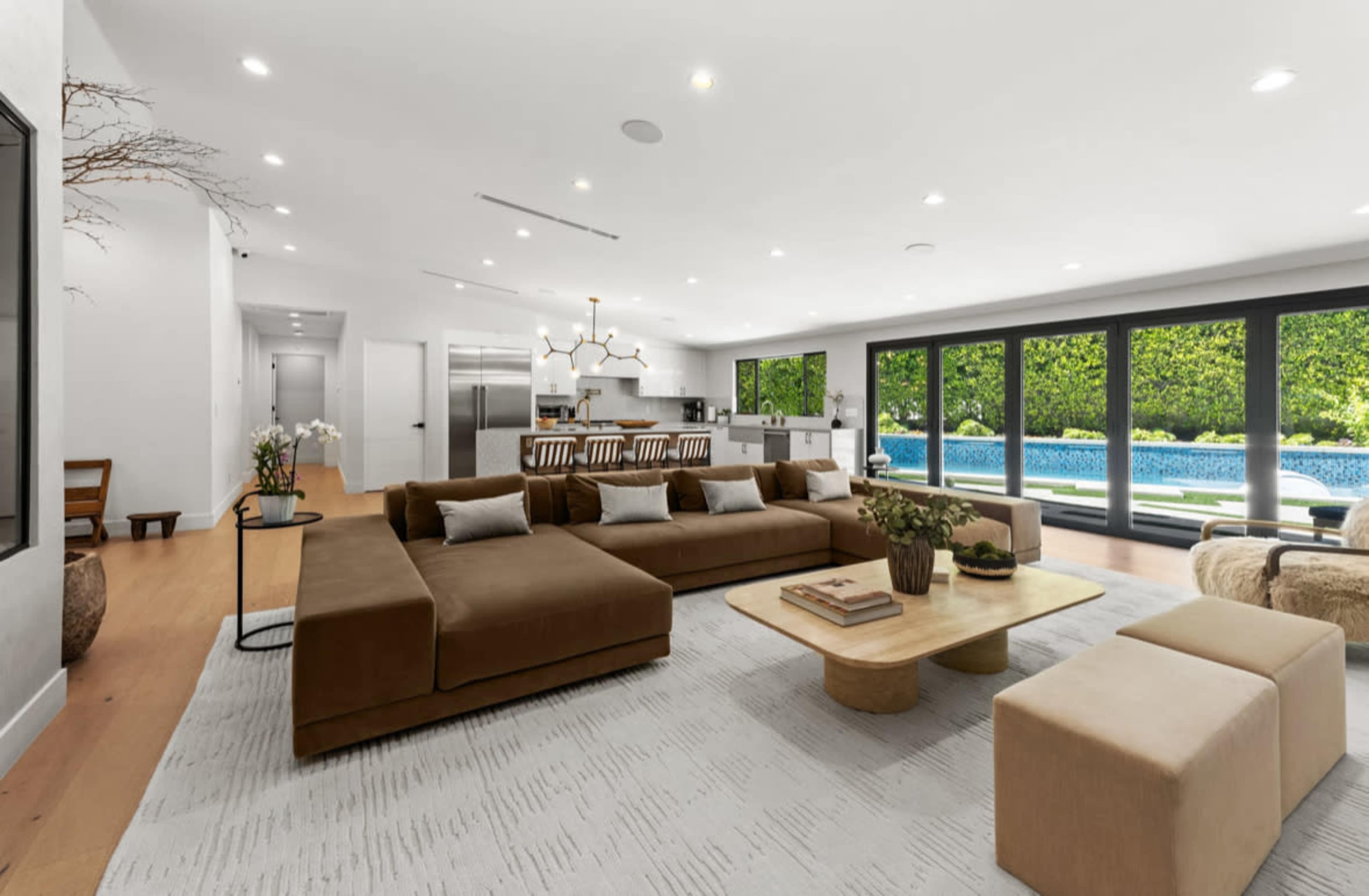 A modern open-concept living room with a large brown sectional sofa, a light coffee table, and a view of a swimming pool through expansive glass doors.
