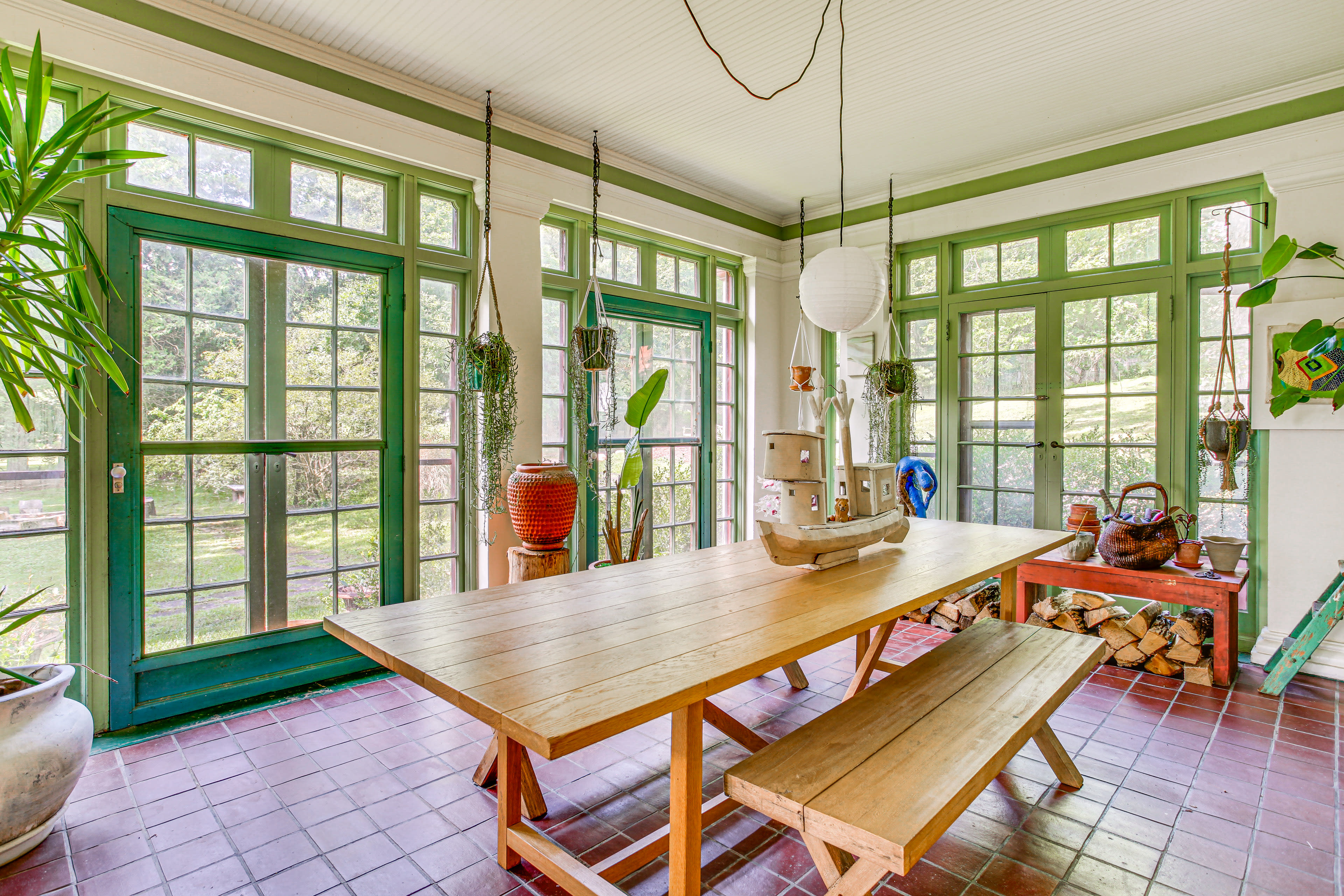 The image shows a sunlit dining area with a large wooden table surrounded by benches, framed by green windows and doors that open to a garden view.