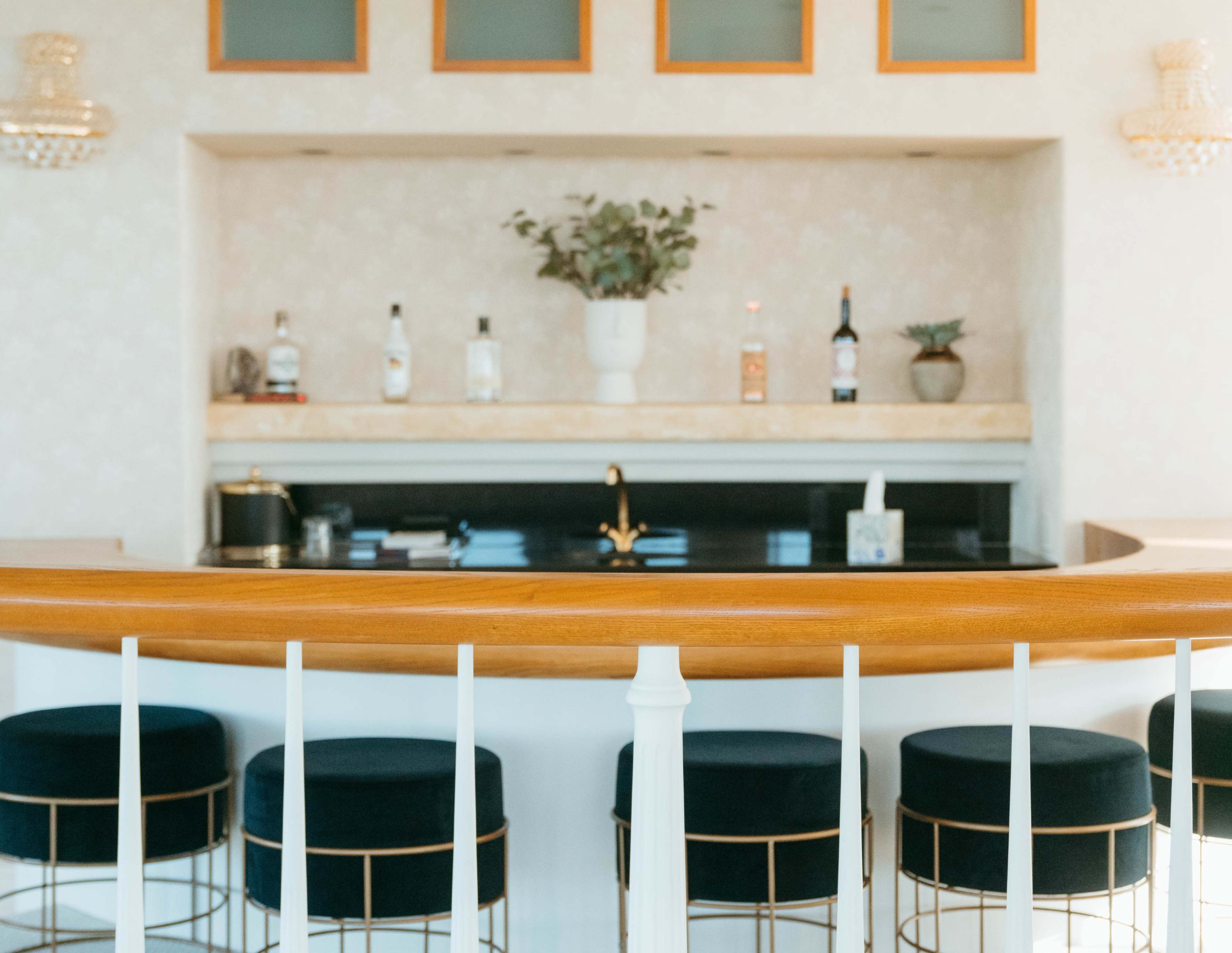 A modern home bar area featuring a marble countertop, several bottles of liquor on display, and upholstered bar stools with gold metal frames.
