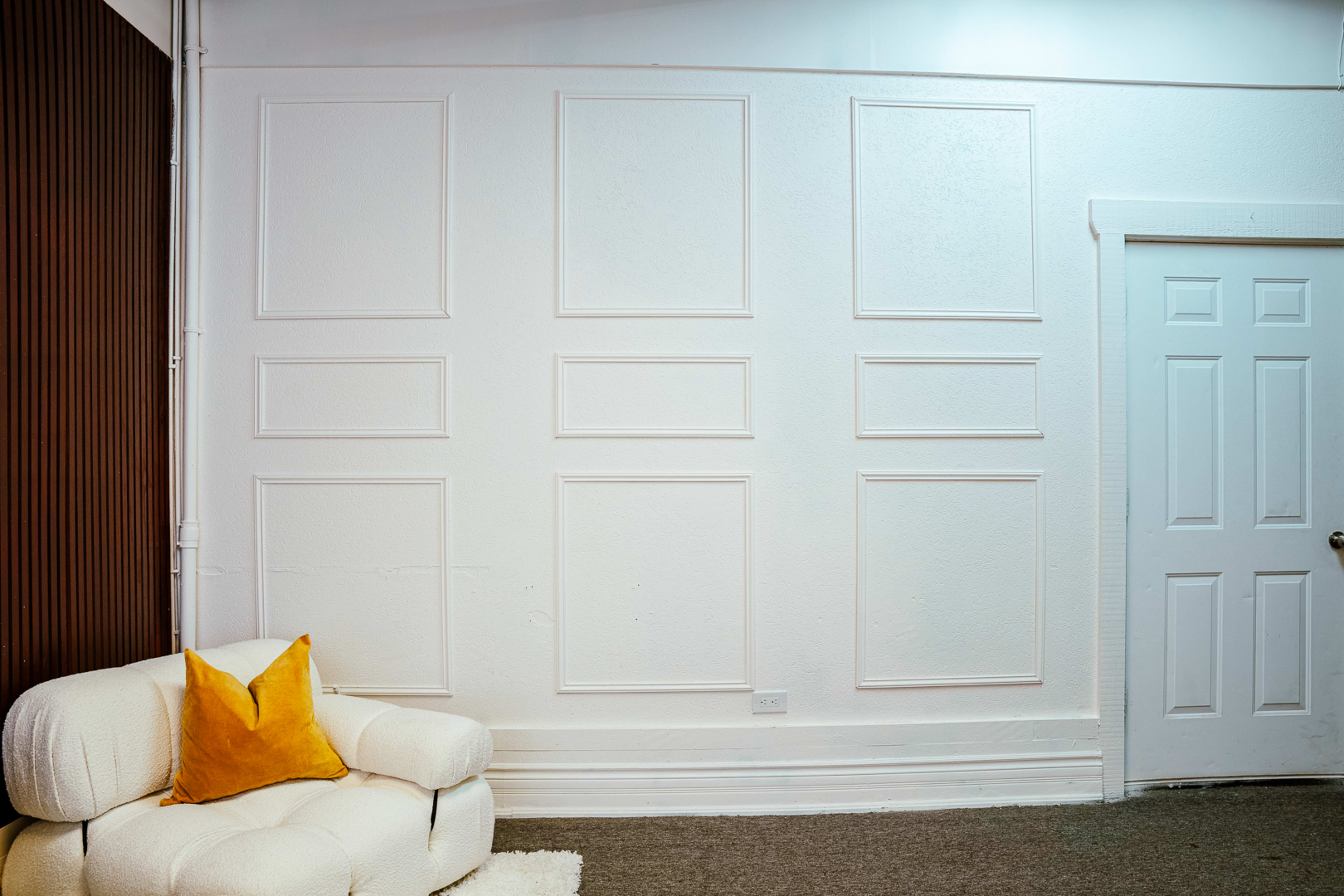 The image shows a corner of a room featuring a textured white wall with decorative moldings, a light-colored couch with an orange pillow, and a white door.