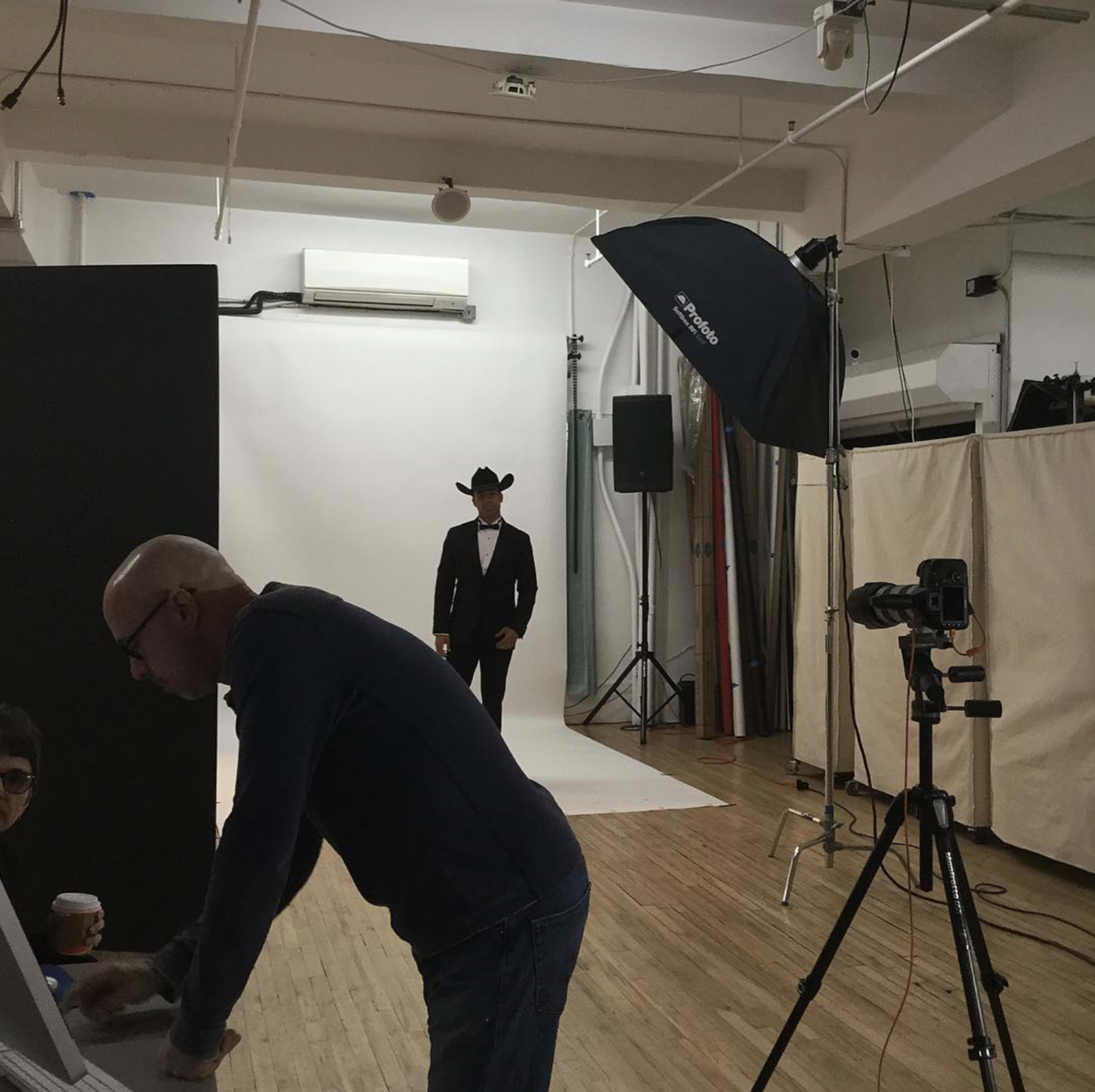 A photographer adjusts equipment in a studio while a model poses in a cowboy hat against a plain backdrop.