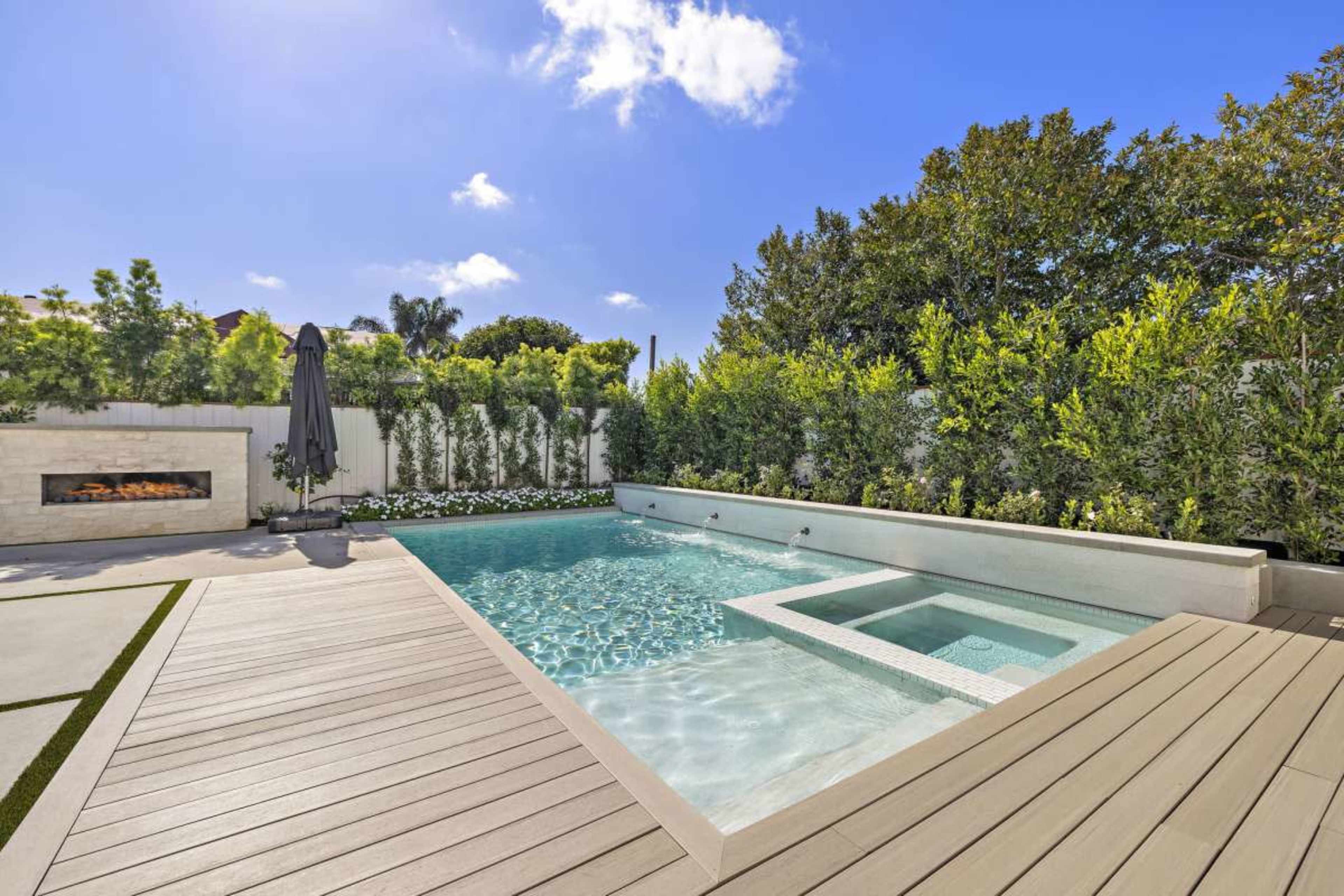 The image shows a modern backyard featuring a swimming pool with a spa, surrounded by neatly arranged greenery and a patio area with composite decking.