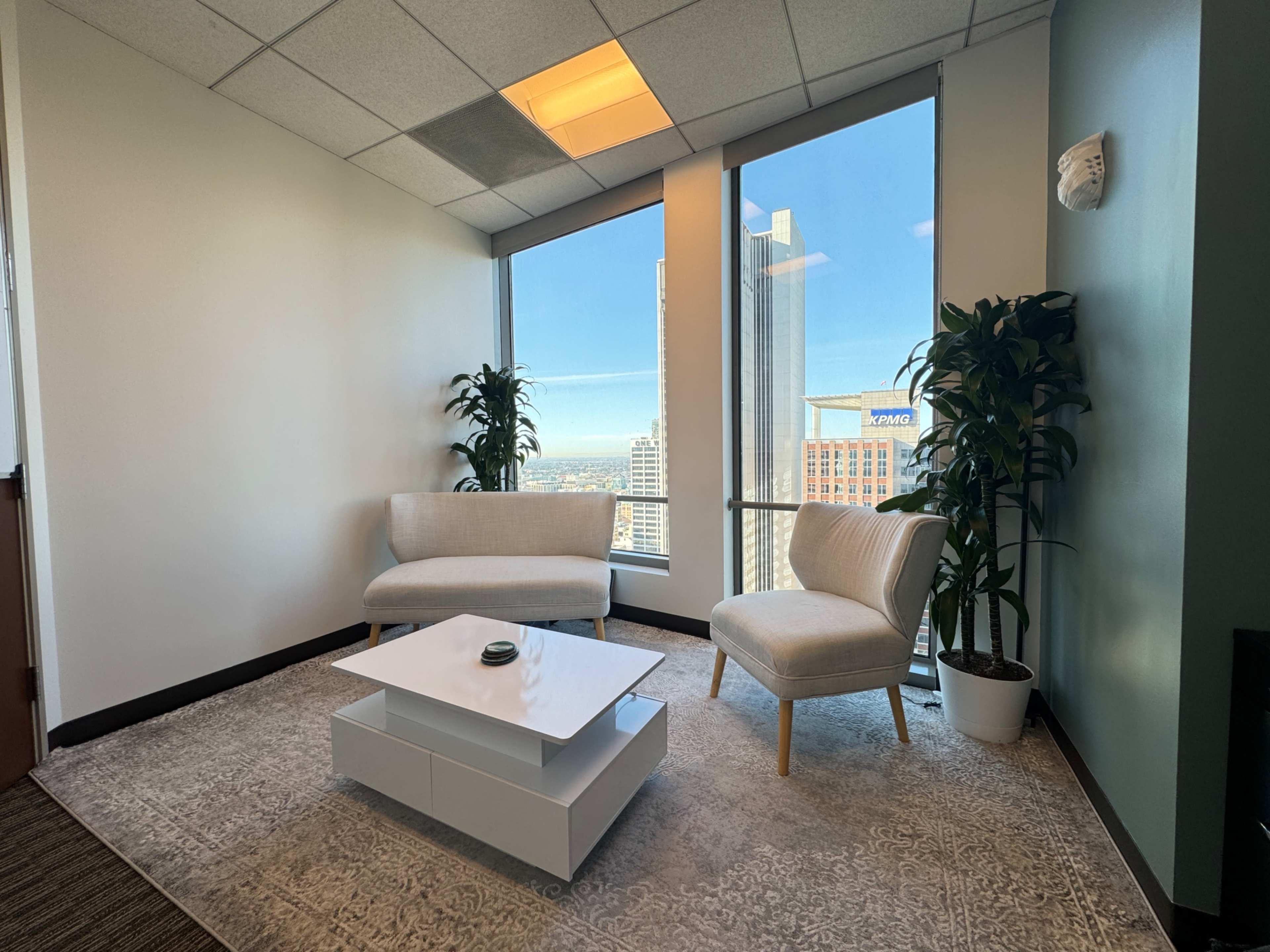 The image shows a modern office waiting area with two chairs, a small white table, and large windows overlooking a cityscape.
