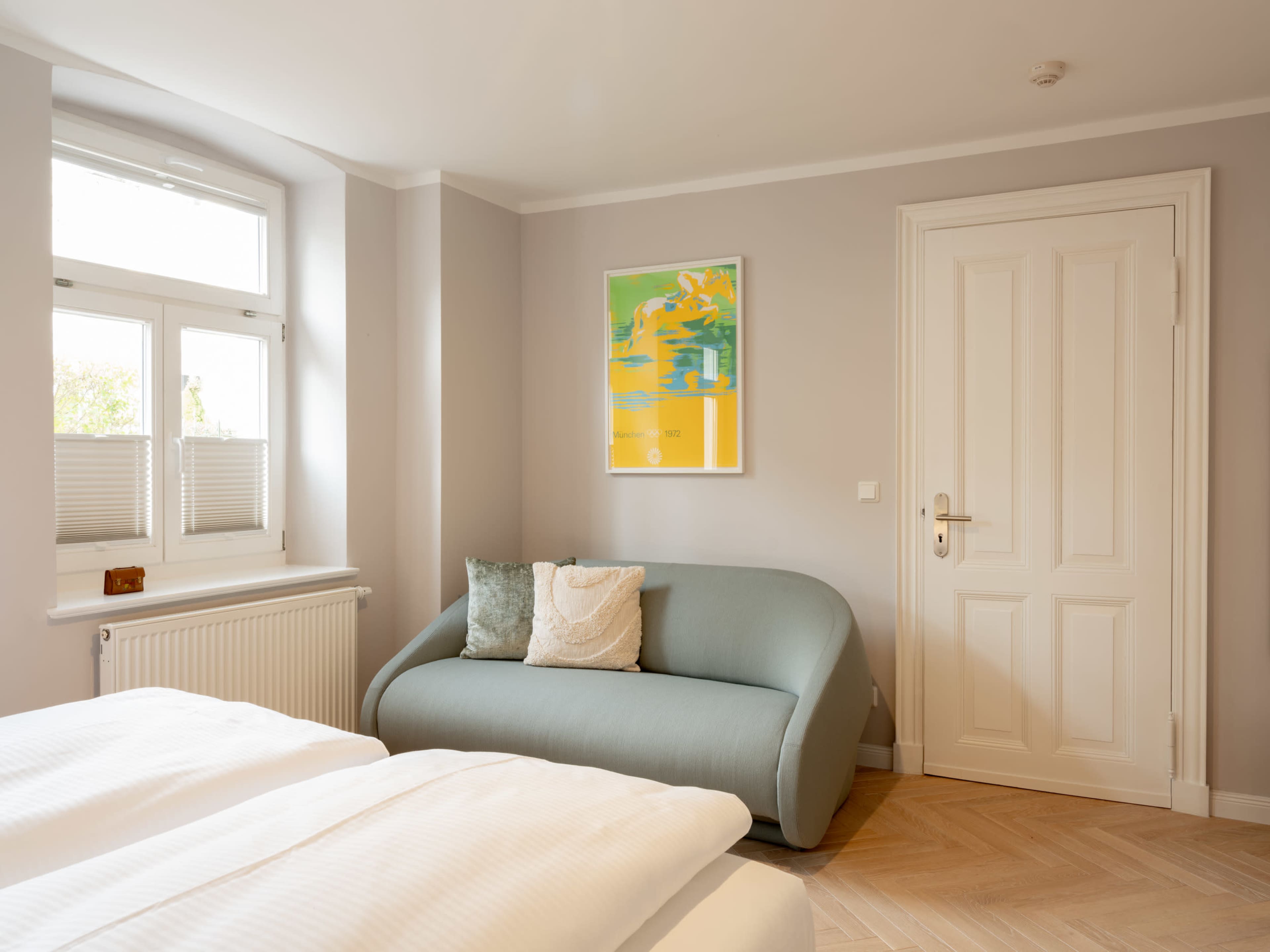 The image shows a light-colored bedroom with a bed, a green sofa, a window with white blinds, and a door.
