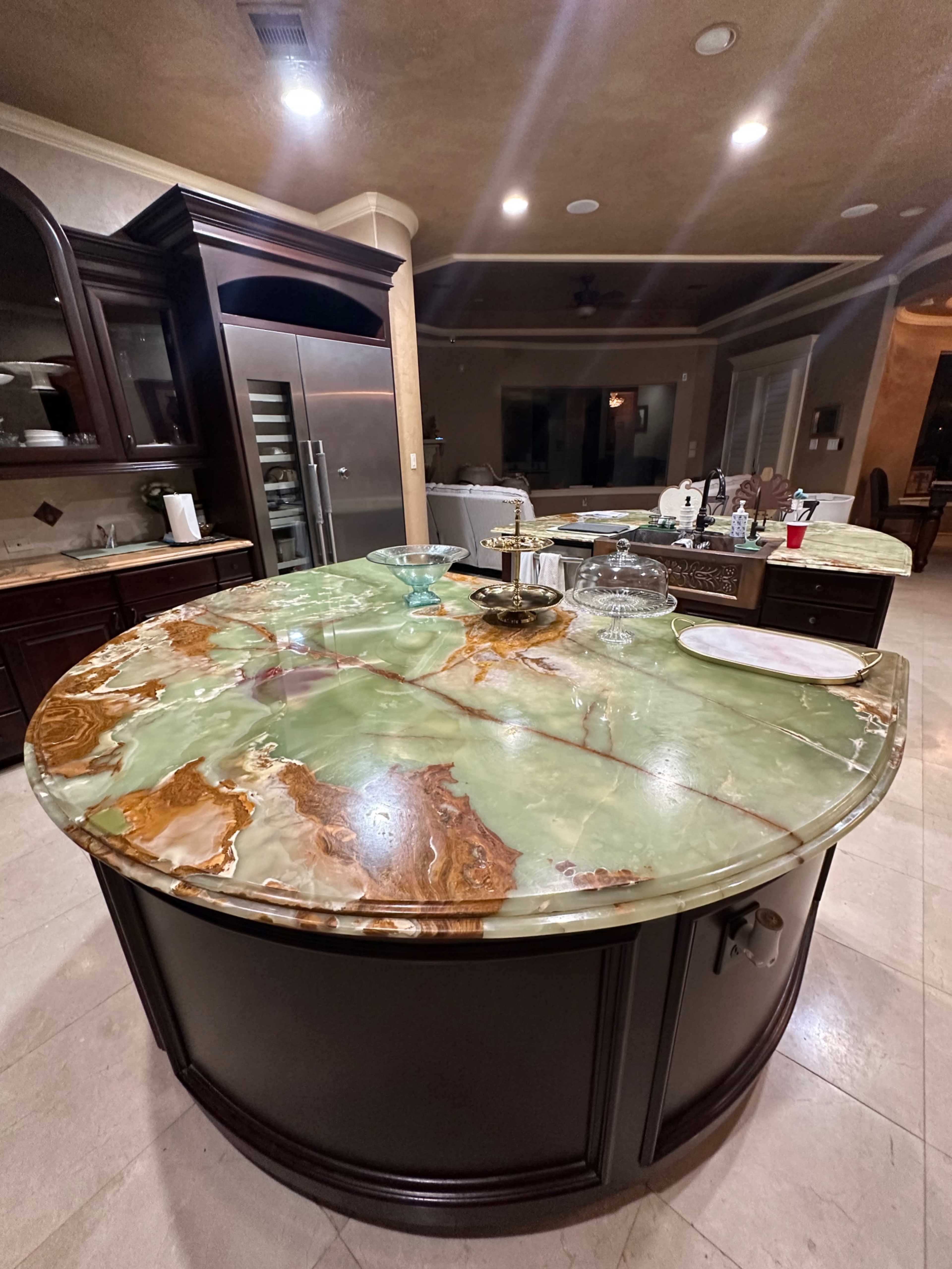 The image shows a large, circular kitchen island with a green and brown stone countertop, surrounded by dark wood cabinetry.