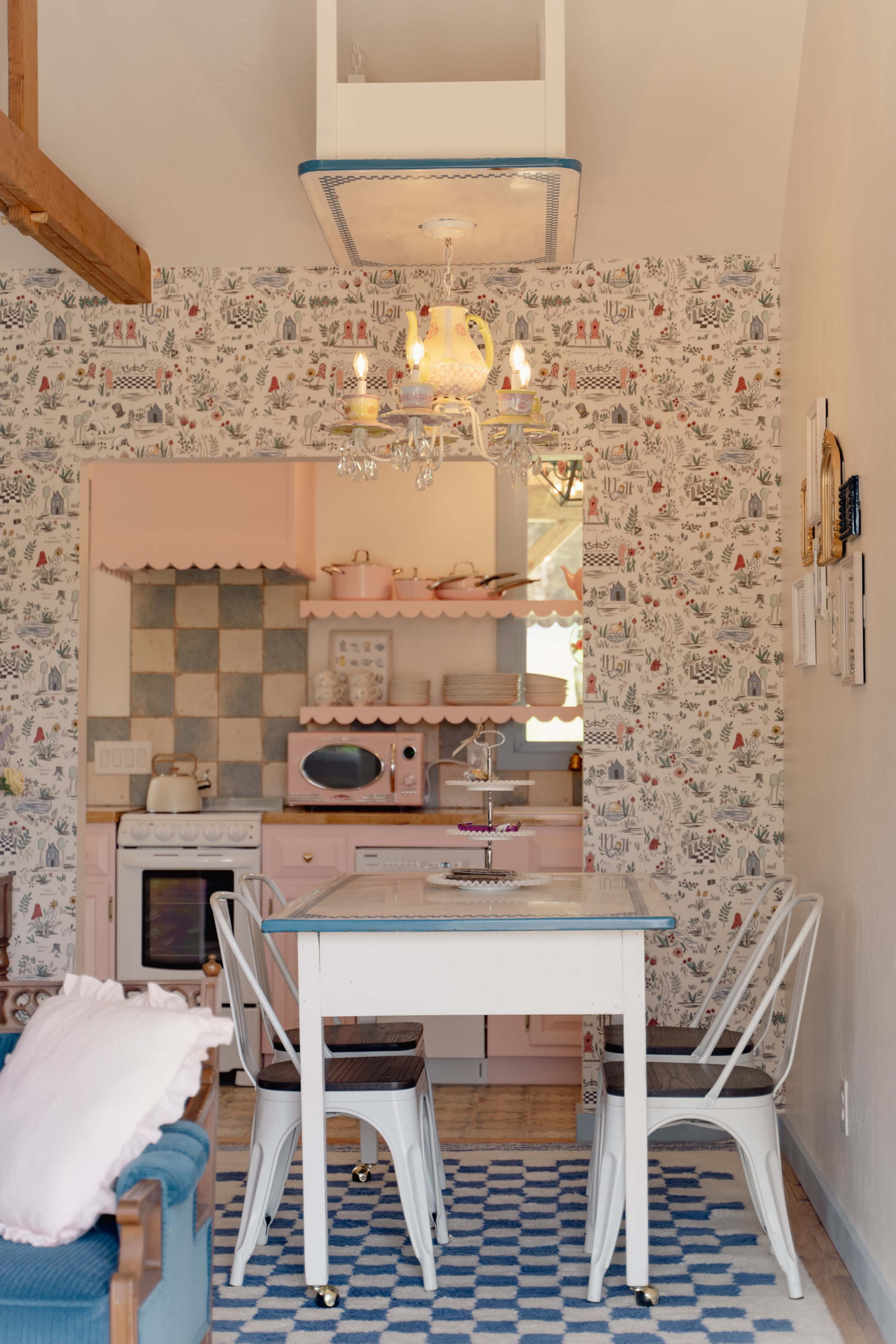 A dining area features a white table and chairs, pink and blue patterned wallpaper, and a small kitchen space with pastel appliances.