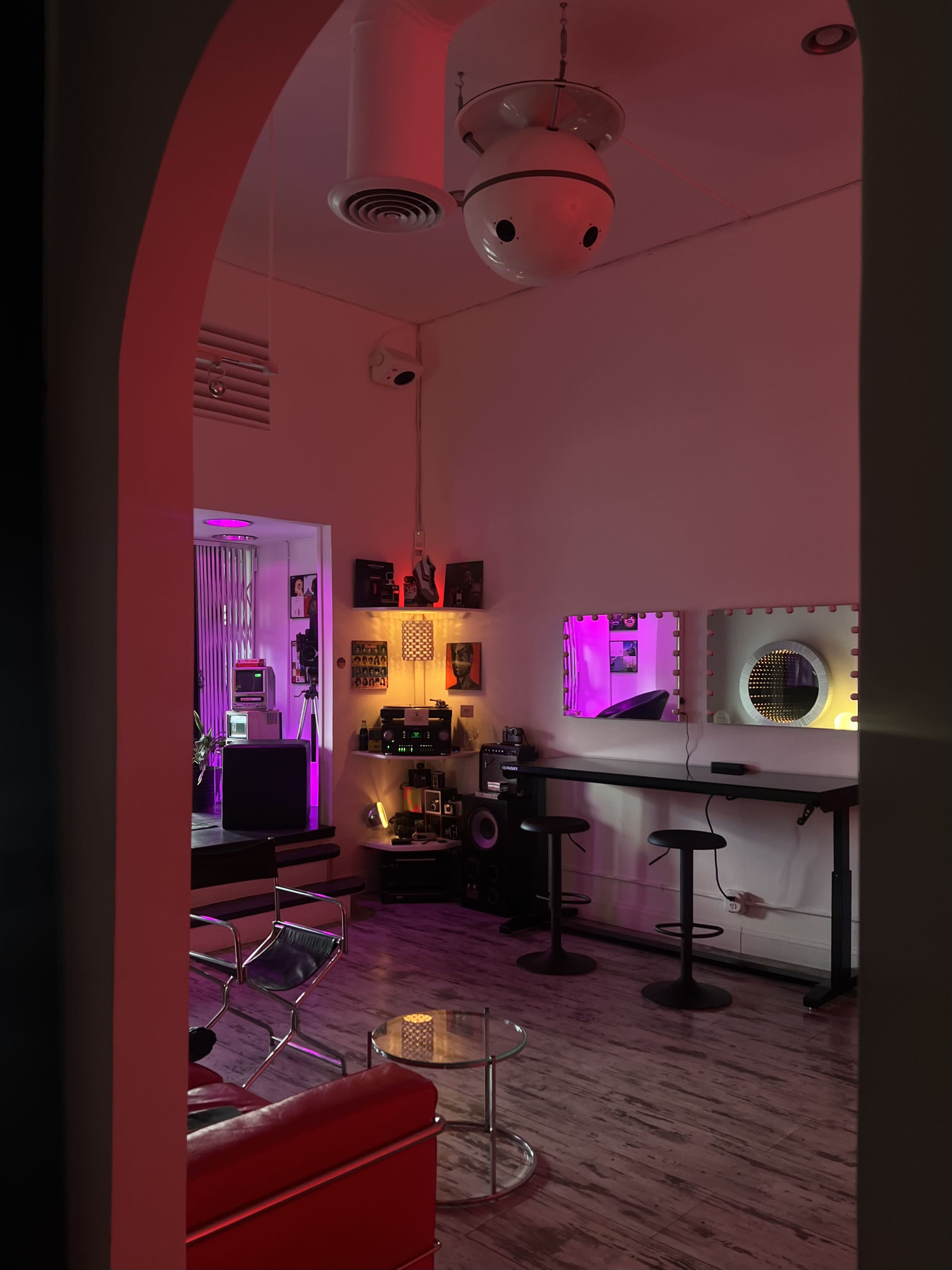 The image shows a stylish studio space with modern furniture, colorful lighting, and various audio equipment.