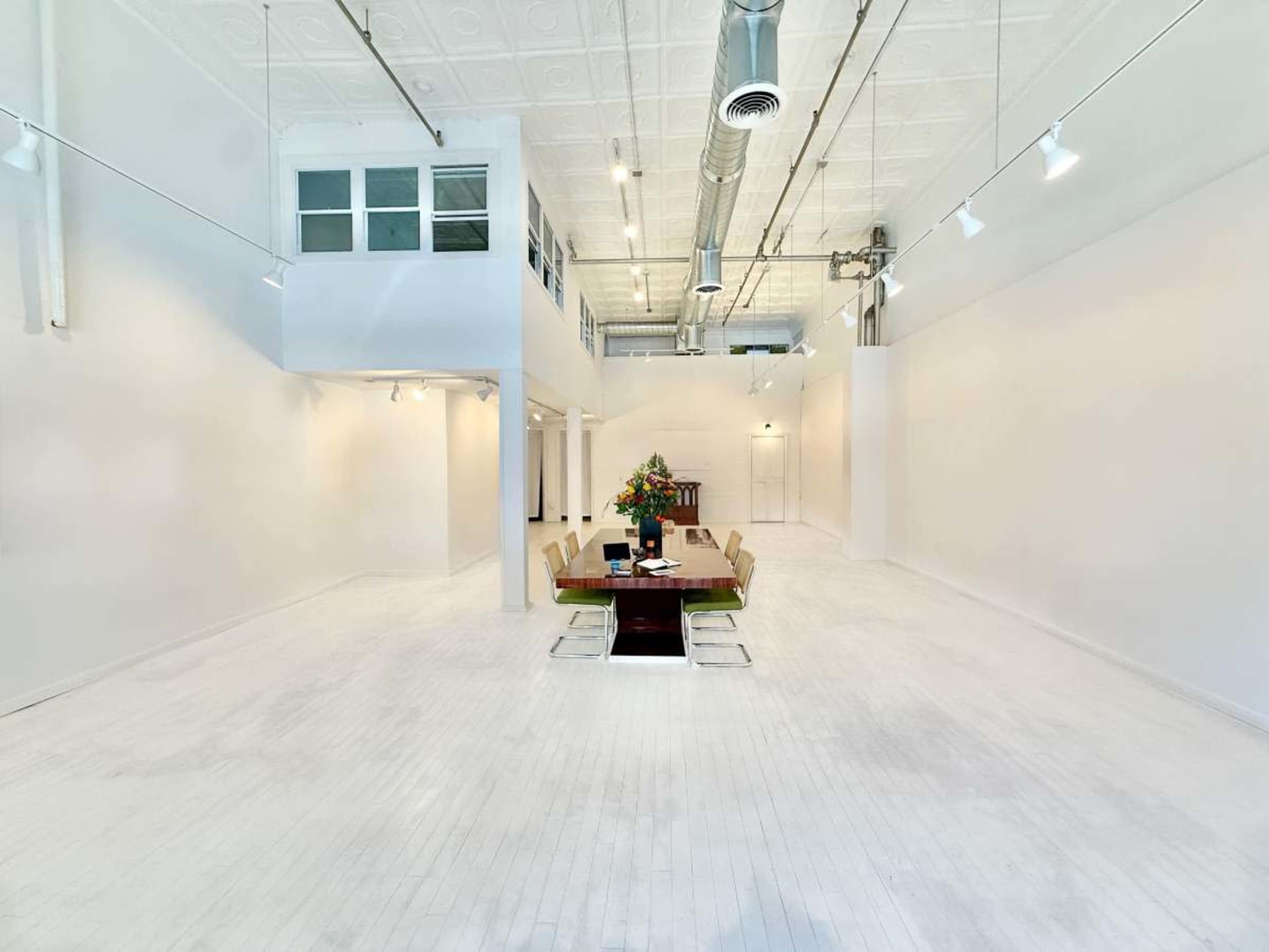 The image shows a spacious, well-lit room with white walls and floors, featuring a central dining table surrounded by chairs and decorative flowers.