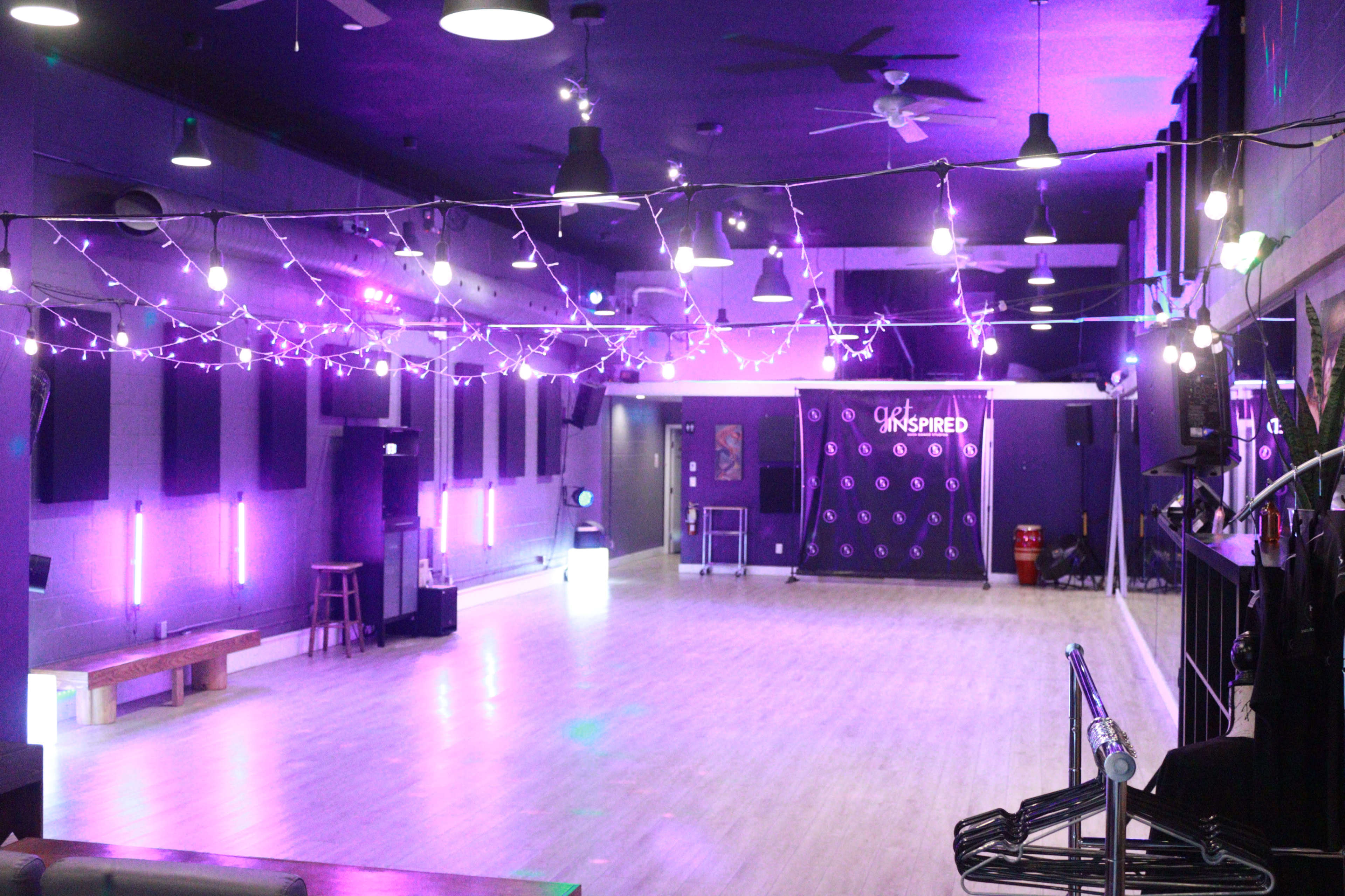 A spacious dance studio with purple lighting, wooden flooring, and string lights overhead.