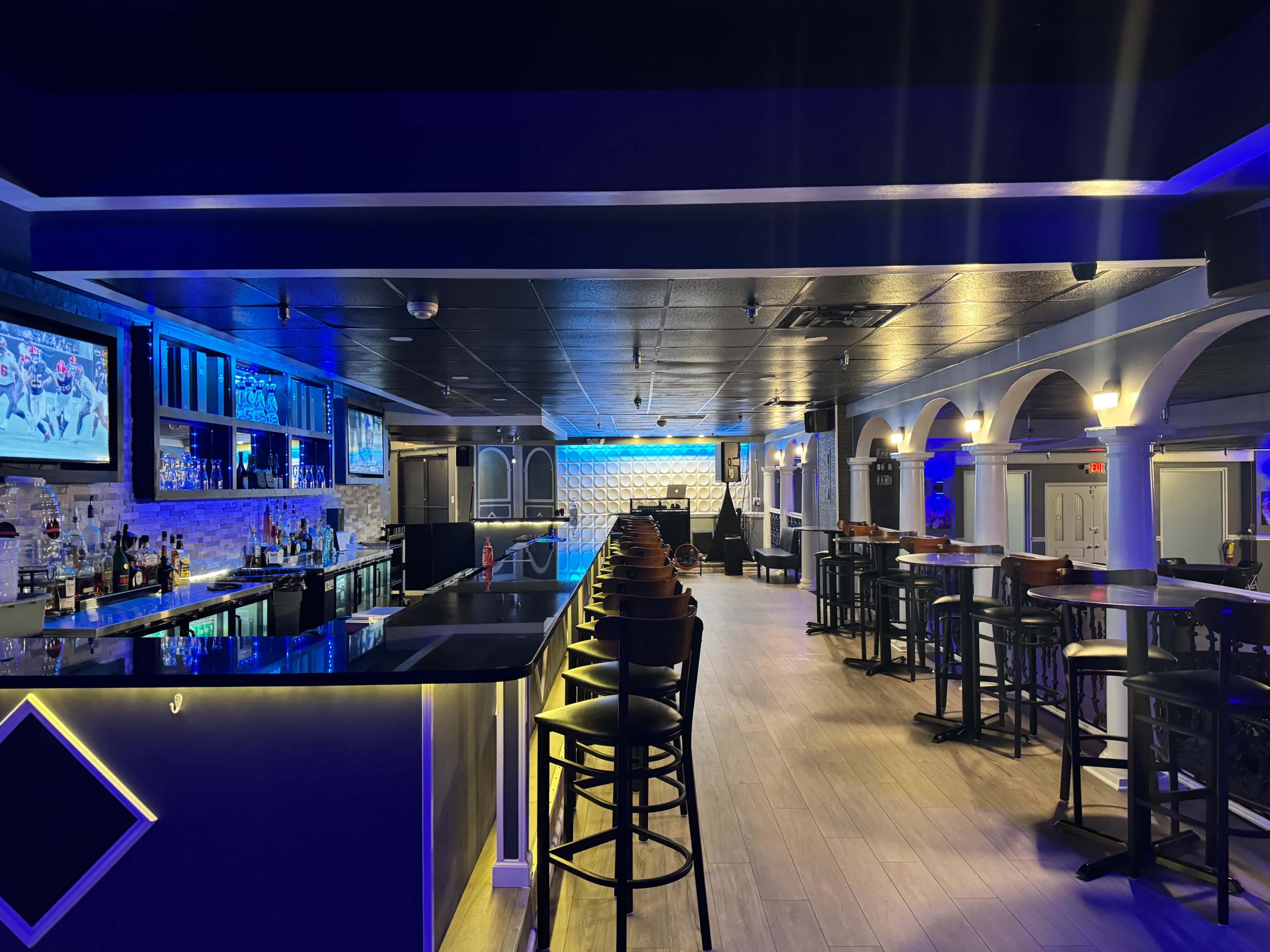 The image shows a modern bar interior featuring a long counter with high stools, illuminated by blue lighting, and multiple televisions mounted on the walls.