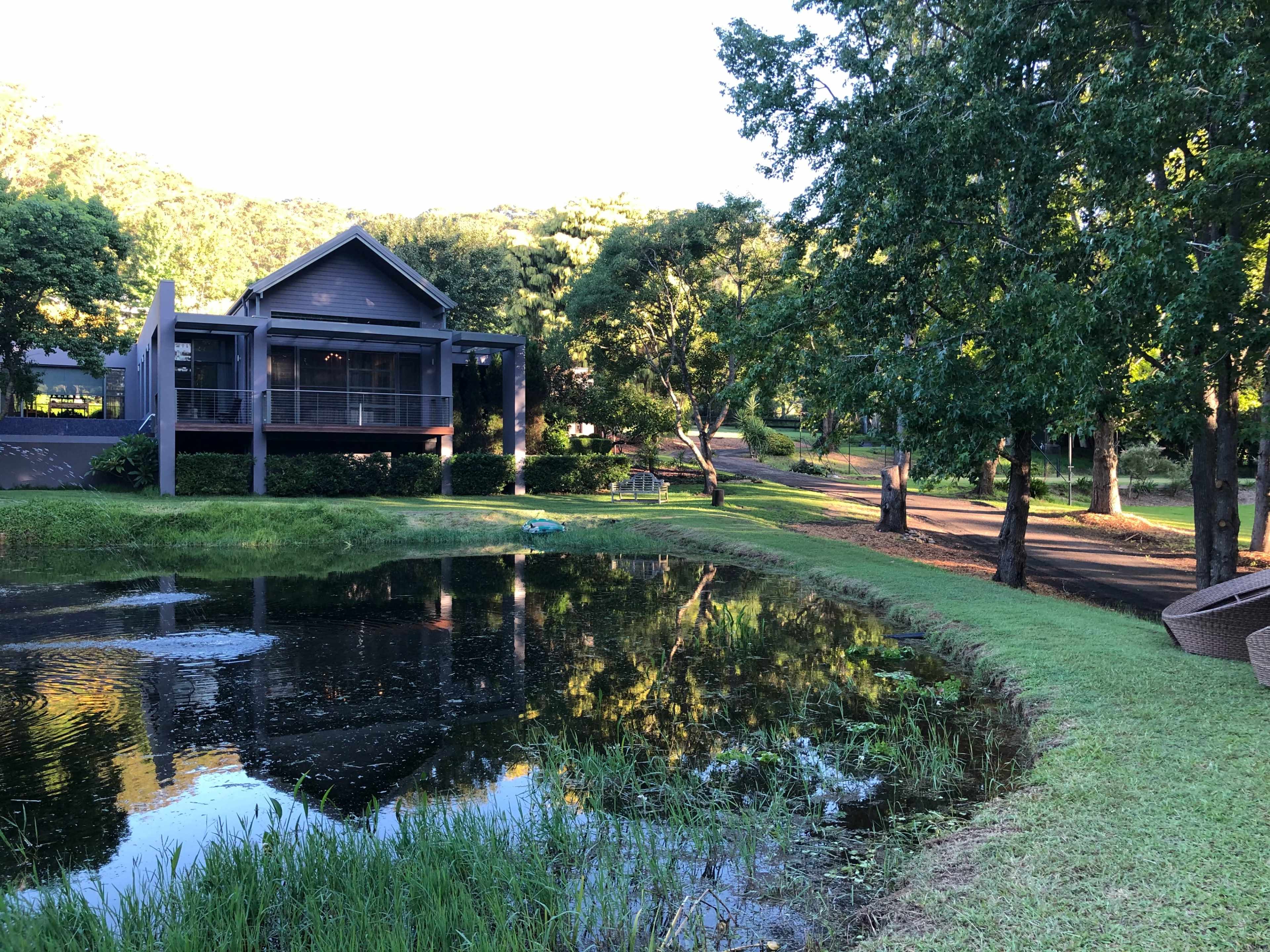 A house with a deck sits beside a pond, surrounded by trees and a grassy area.
