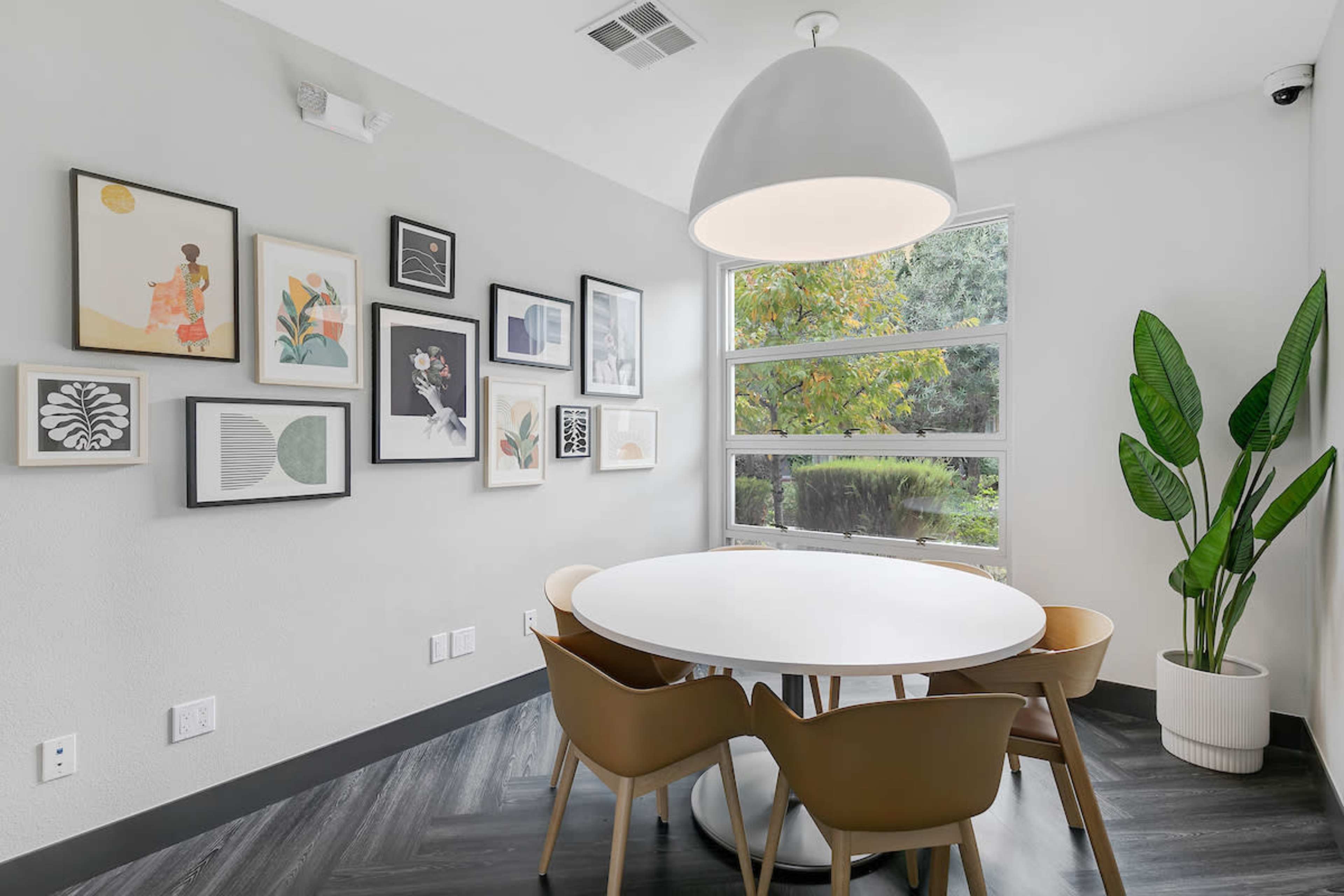 A round white table with four brown chairs is situated in a room featuring large windows and a gallery wall of framed artwork.