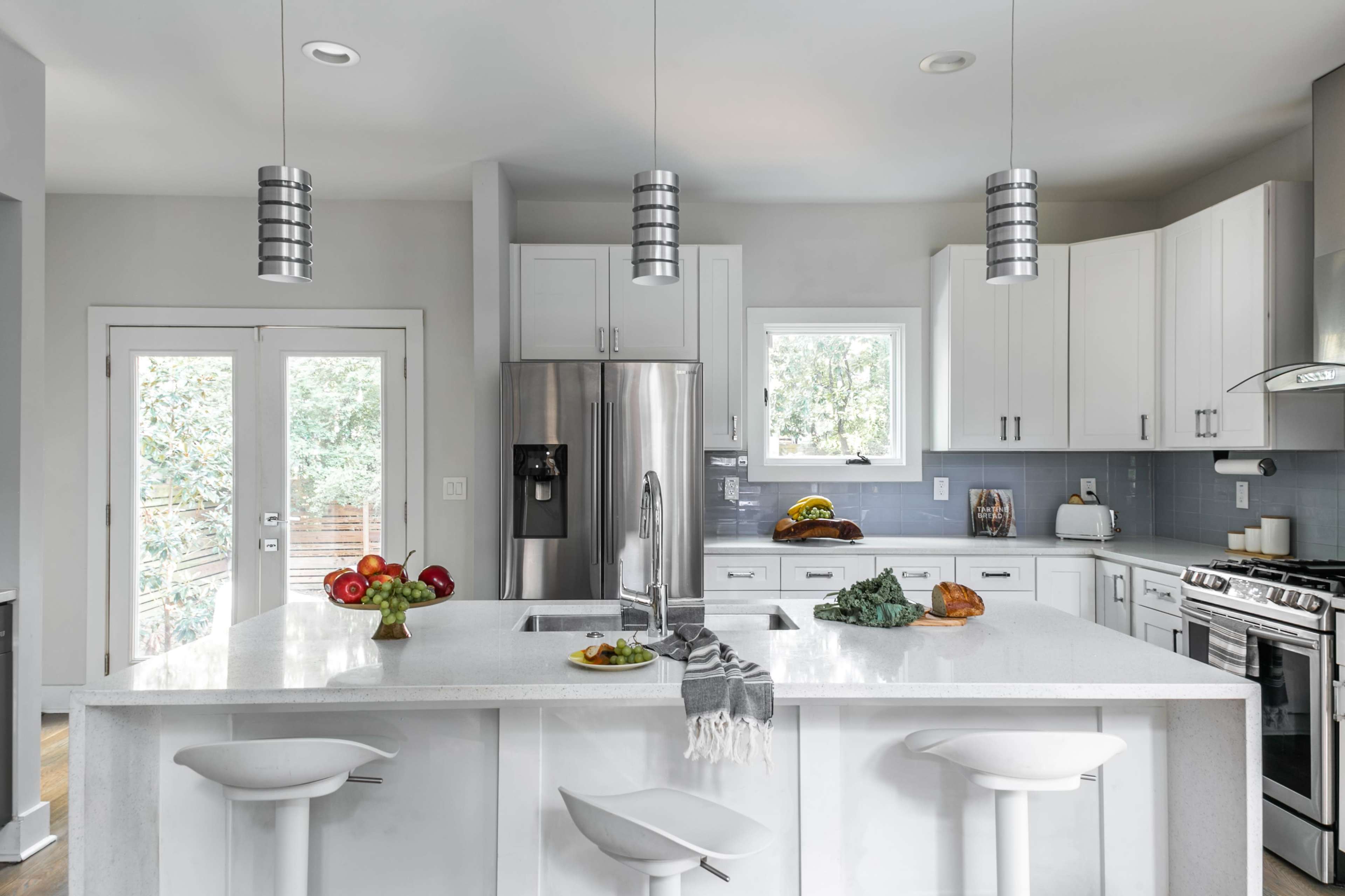 A modern kitchen features a large island with bar stools, stainless steel appliances, and white cabinetry, illuminated by pendant lights.
