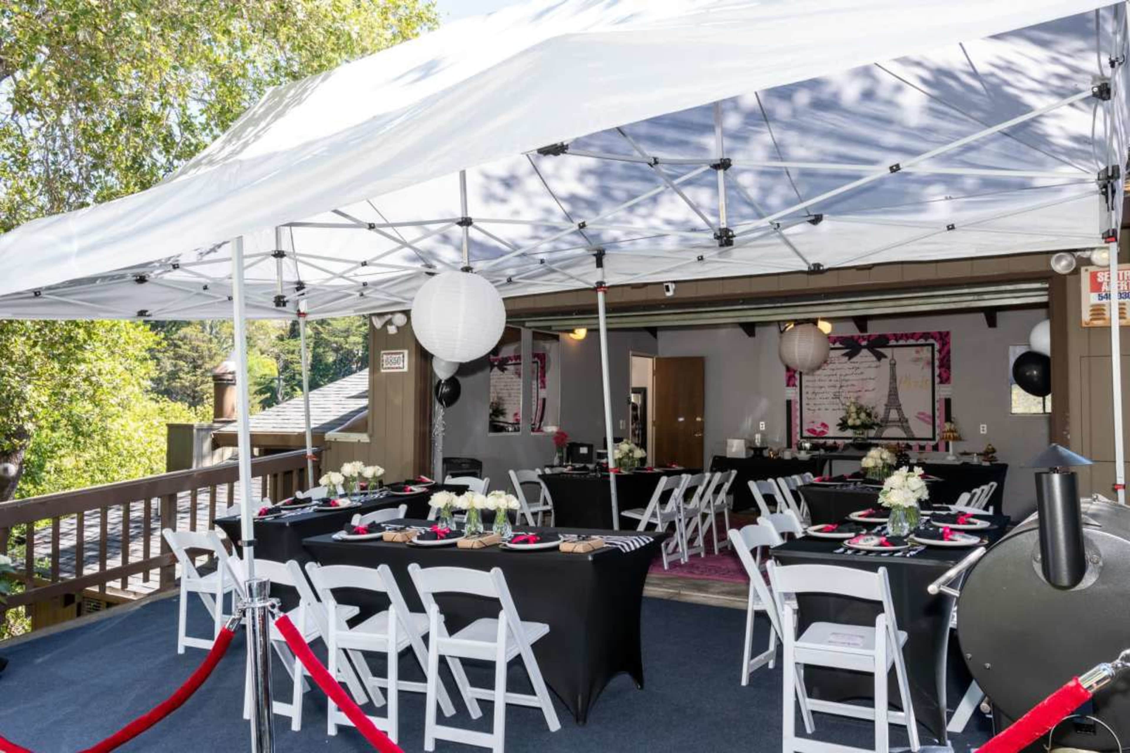 A covered outdoor seating area is set up for an event with black and white tables, white flowers, and elegant decorations.