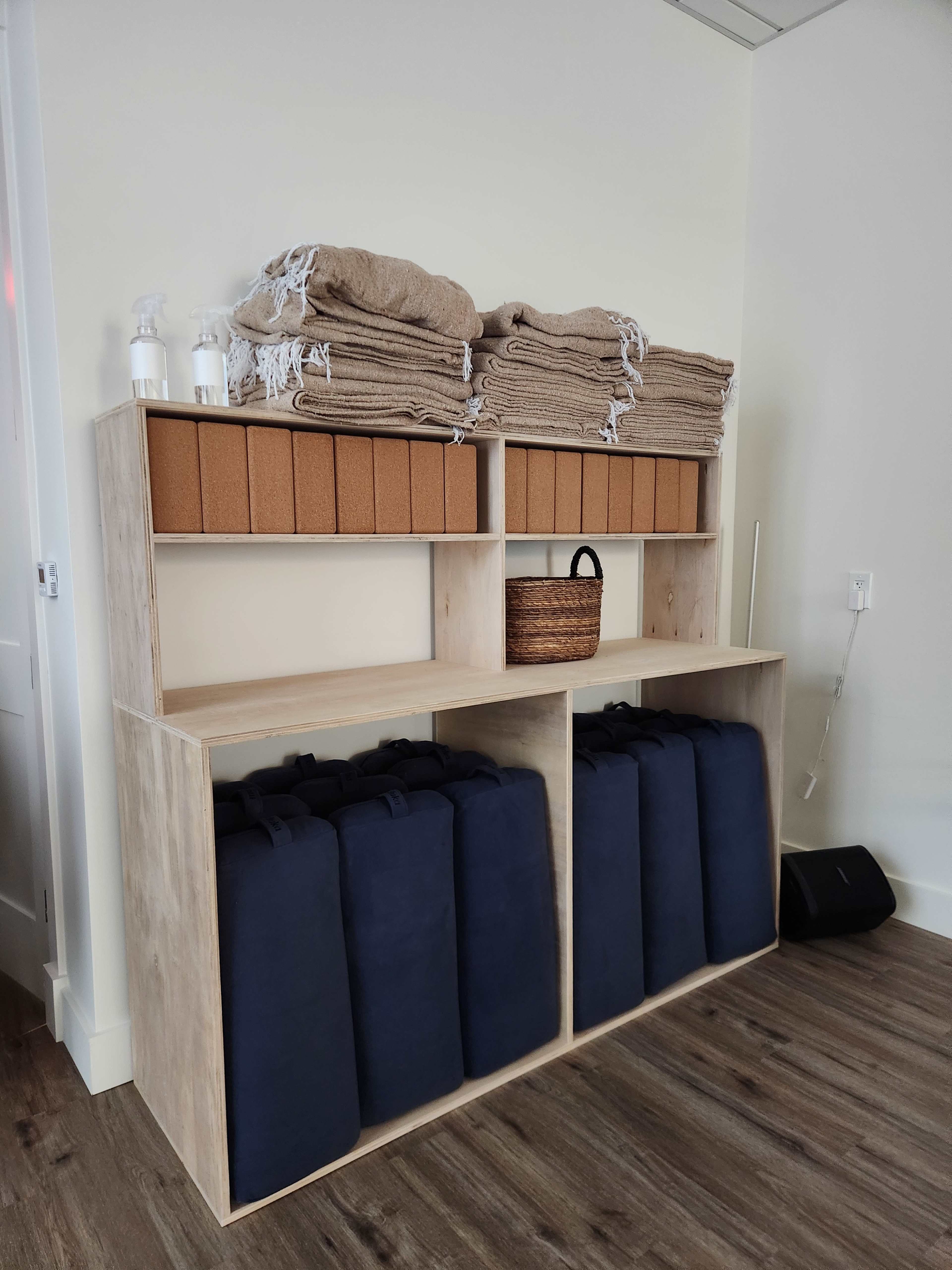 The image shows a wooden shelving unit with stacked towels on top, a woven basket, and several blue padded mats neatly arranged on the bottom.