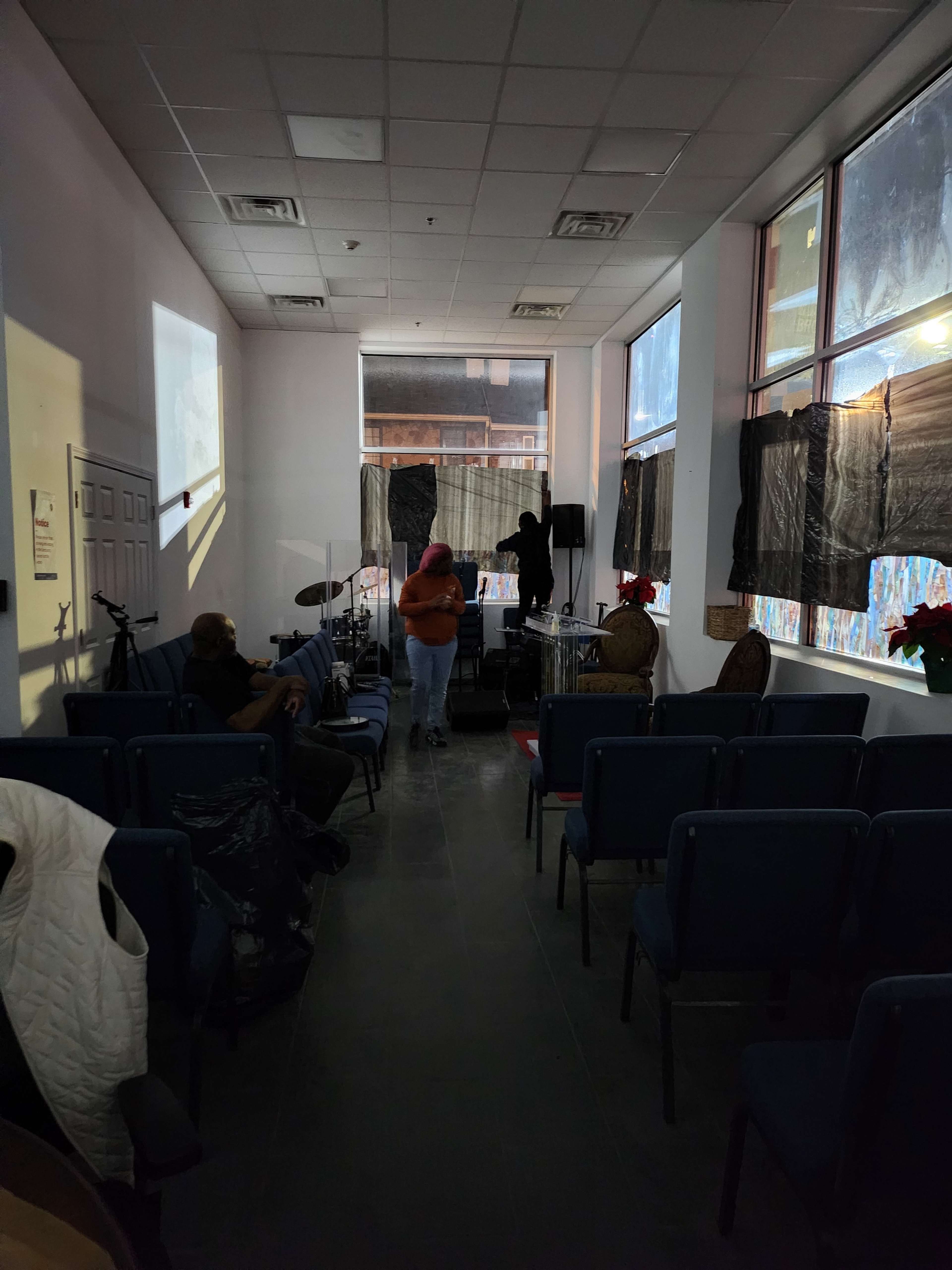 A sparsely furnished room with blue chairs, a stage setup at the front, and large windows covered with dark fabric.