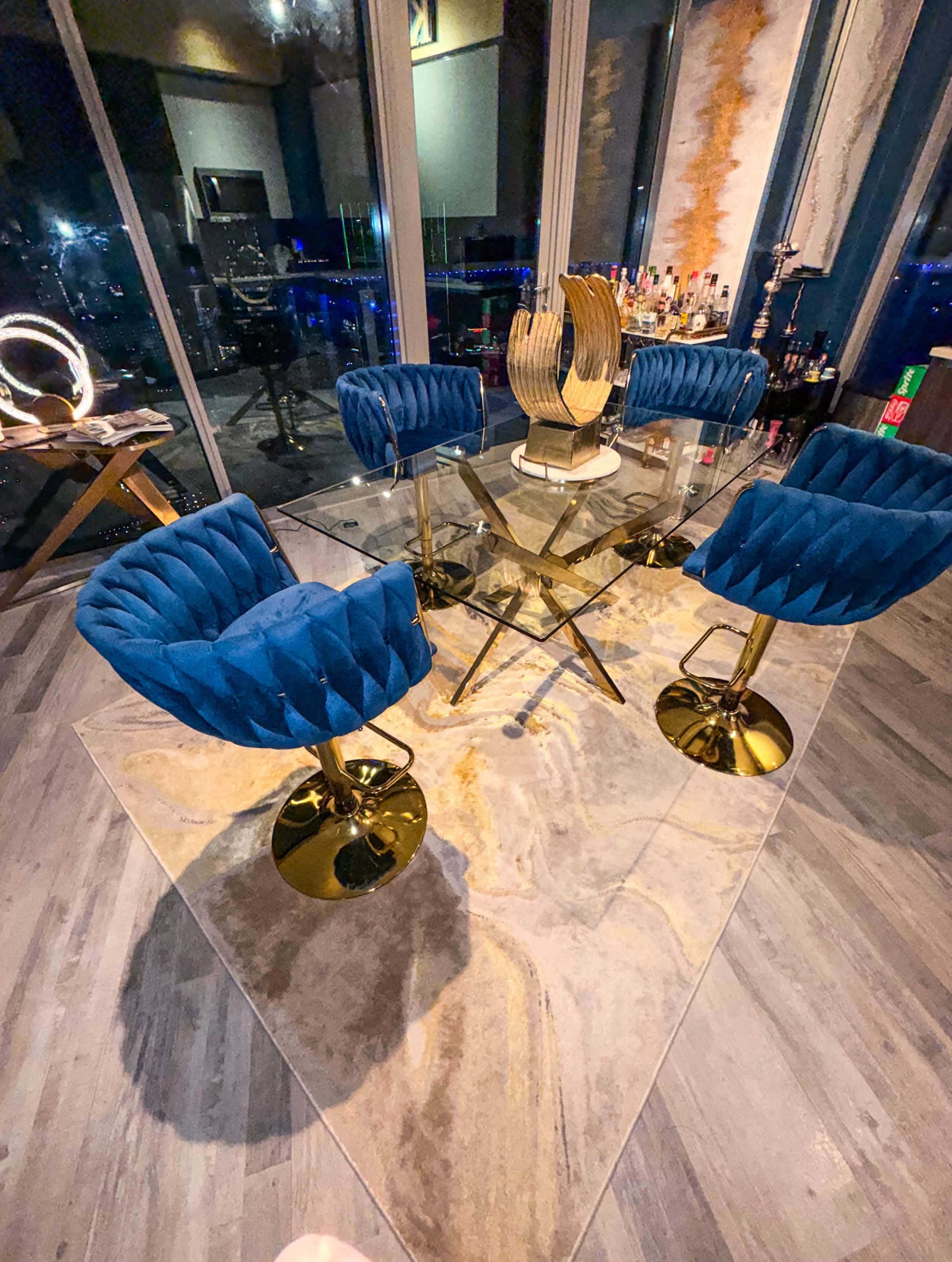 A glass table is surrounded by four blue velvet chairs with gold bases, set on a patterned rug in a well-lit room with a city view.