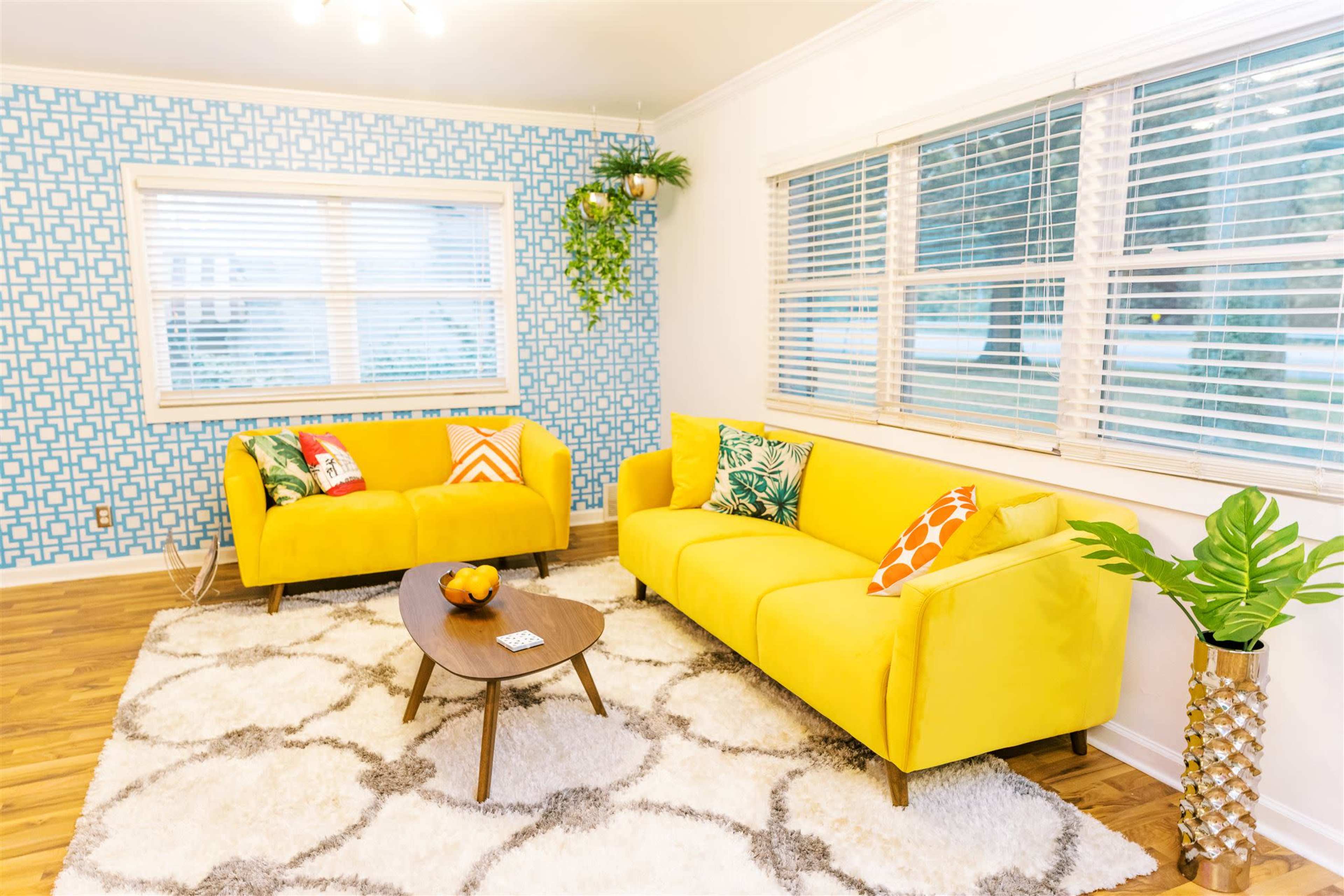 A brightly colored living room featuring yellow sofas, patterned cushions, a wooden coffee table, and a large area rug on a hardwood floor, with a patterned blue wall and large windows.