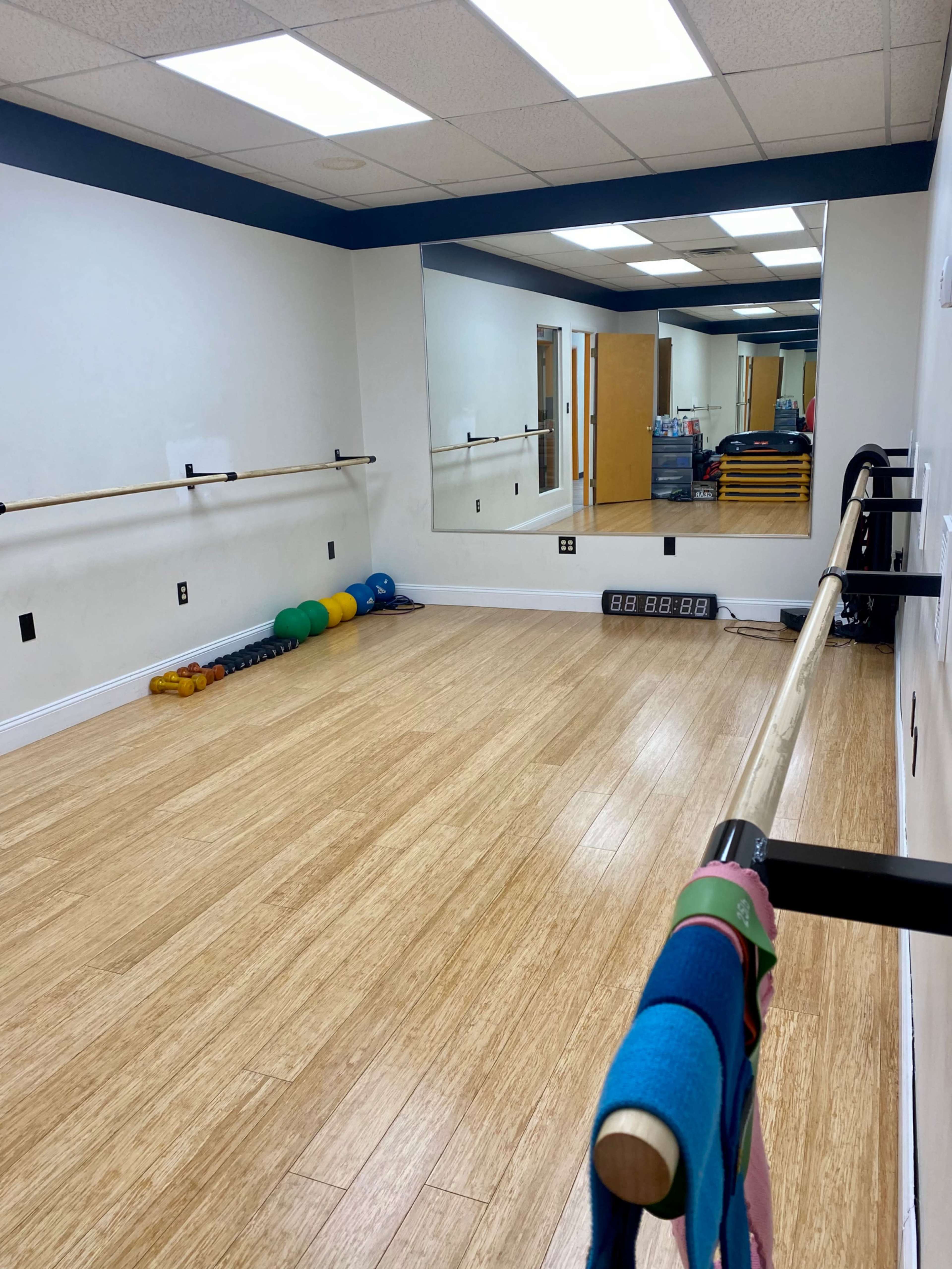 The image shows a fitness studio with wooden flooring, wall-mounted ballet barres, a large mirror, and exercise equipment arranged neatly along the wall.