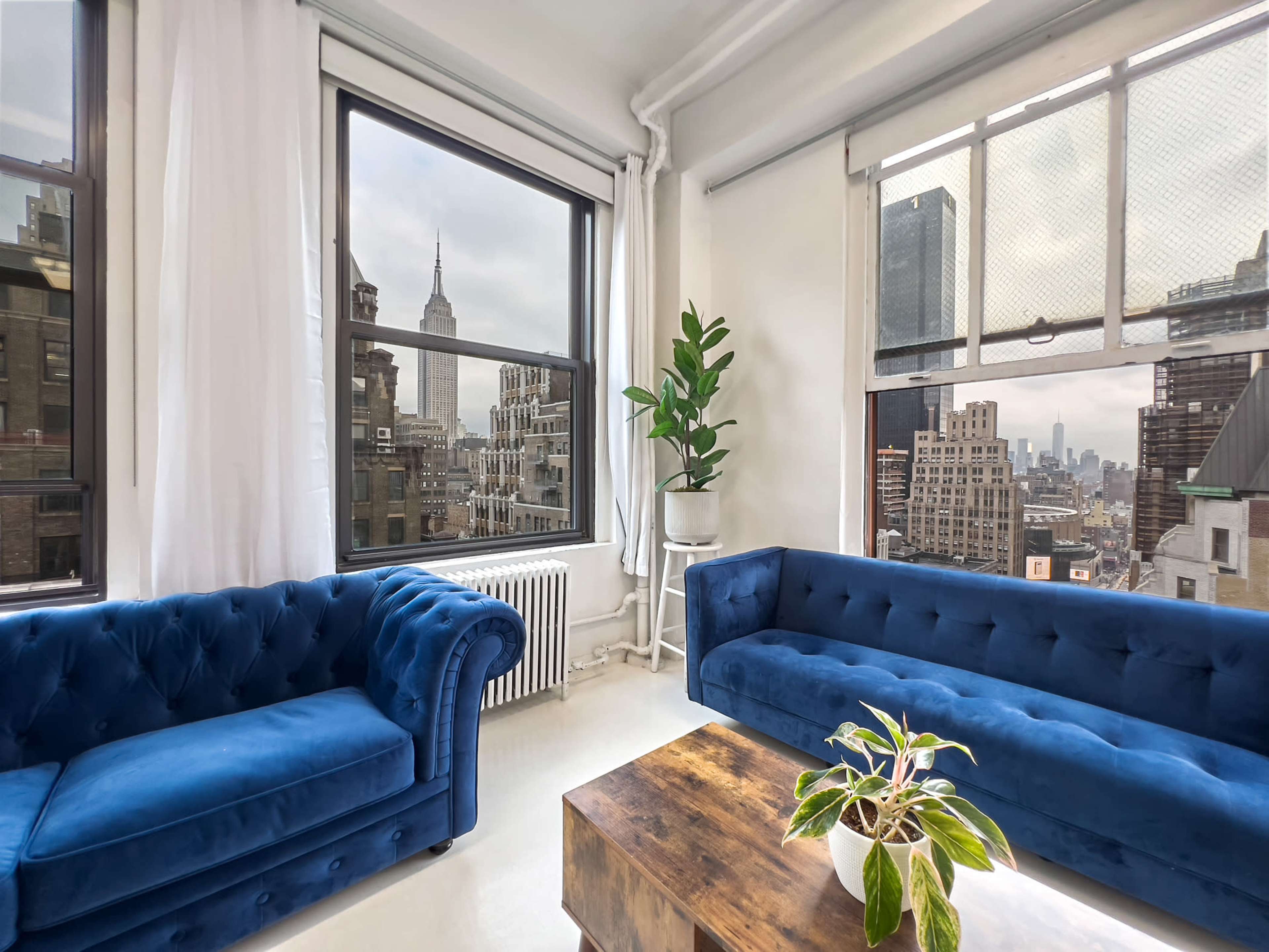 A cozy room with two blue velvet couches, a wooden coffee table, and a large window offering a view of the Empire State Building and city skyline.