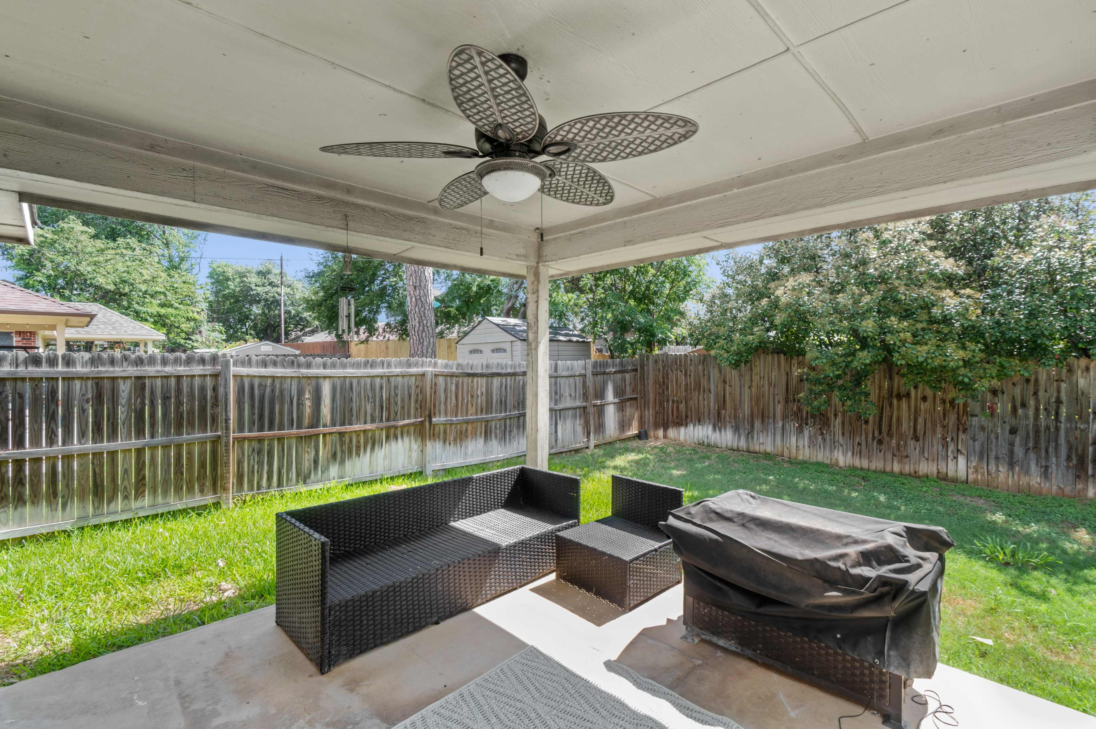 A covered patio features a rattan seating area and a grill, surrounded by a fenced backyard with greenery.
