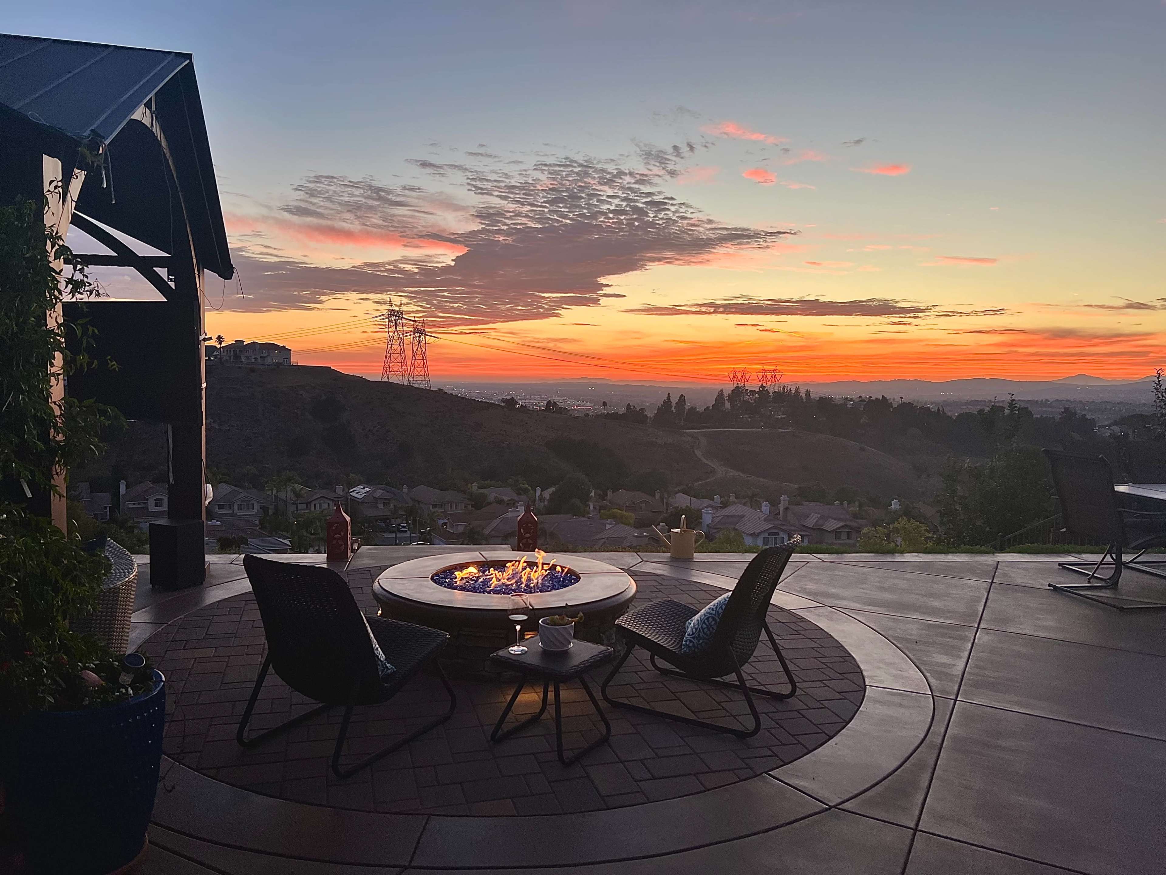 A circular fire pit surrounded by chairs overlooks a hilly landscape at sunset, with power lines and scattered houses in the background.