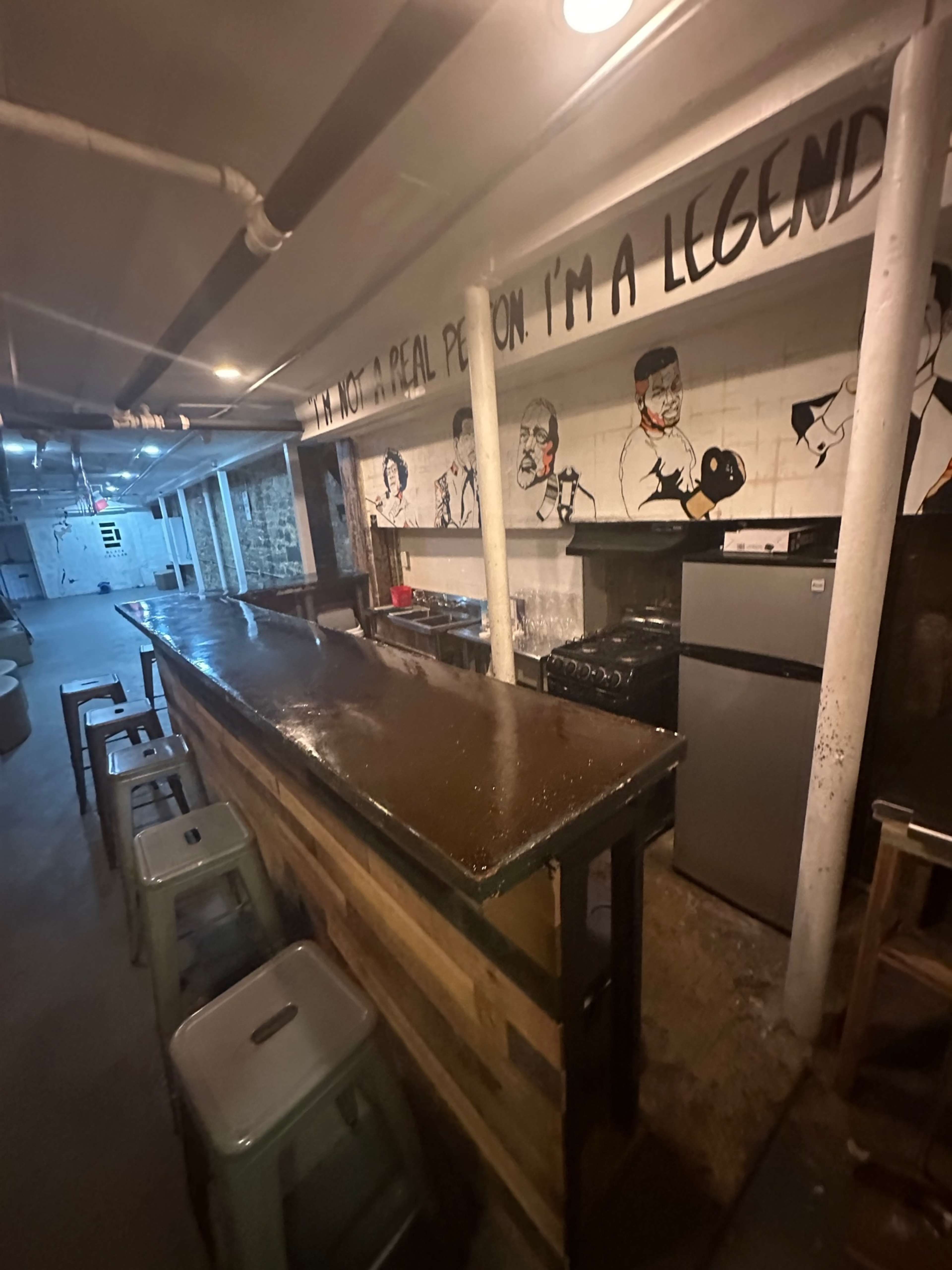The image shows a dimly lit bar area with a wooden counter, metal stools, and murals of boxers on the walls.