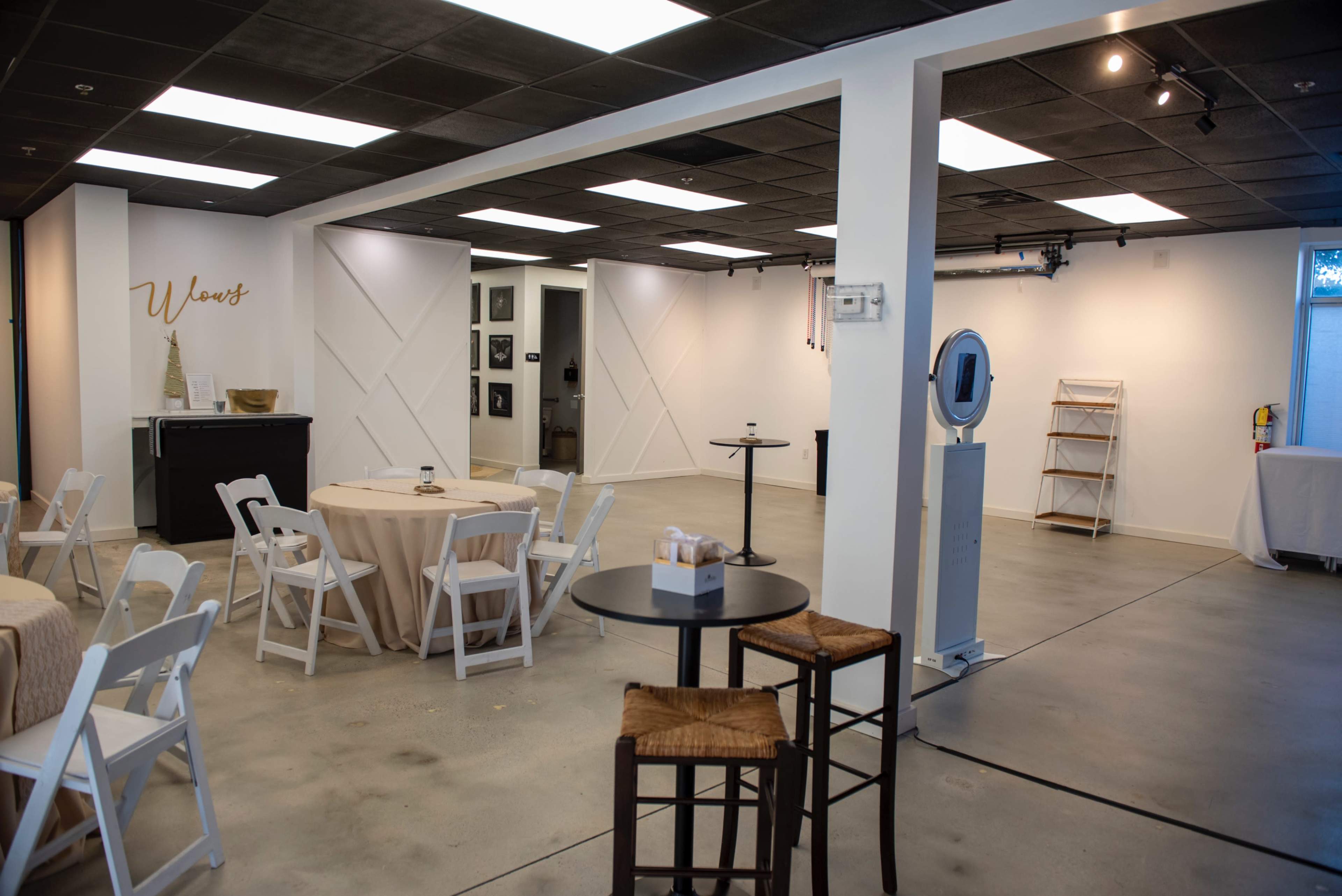 The image shows a spacious, modern event space with tables set up, a bar area, and display shelves against white walls.