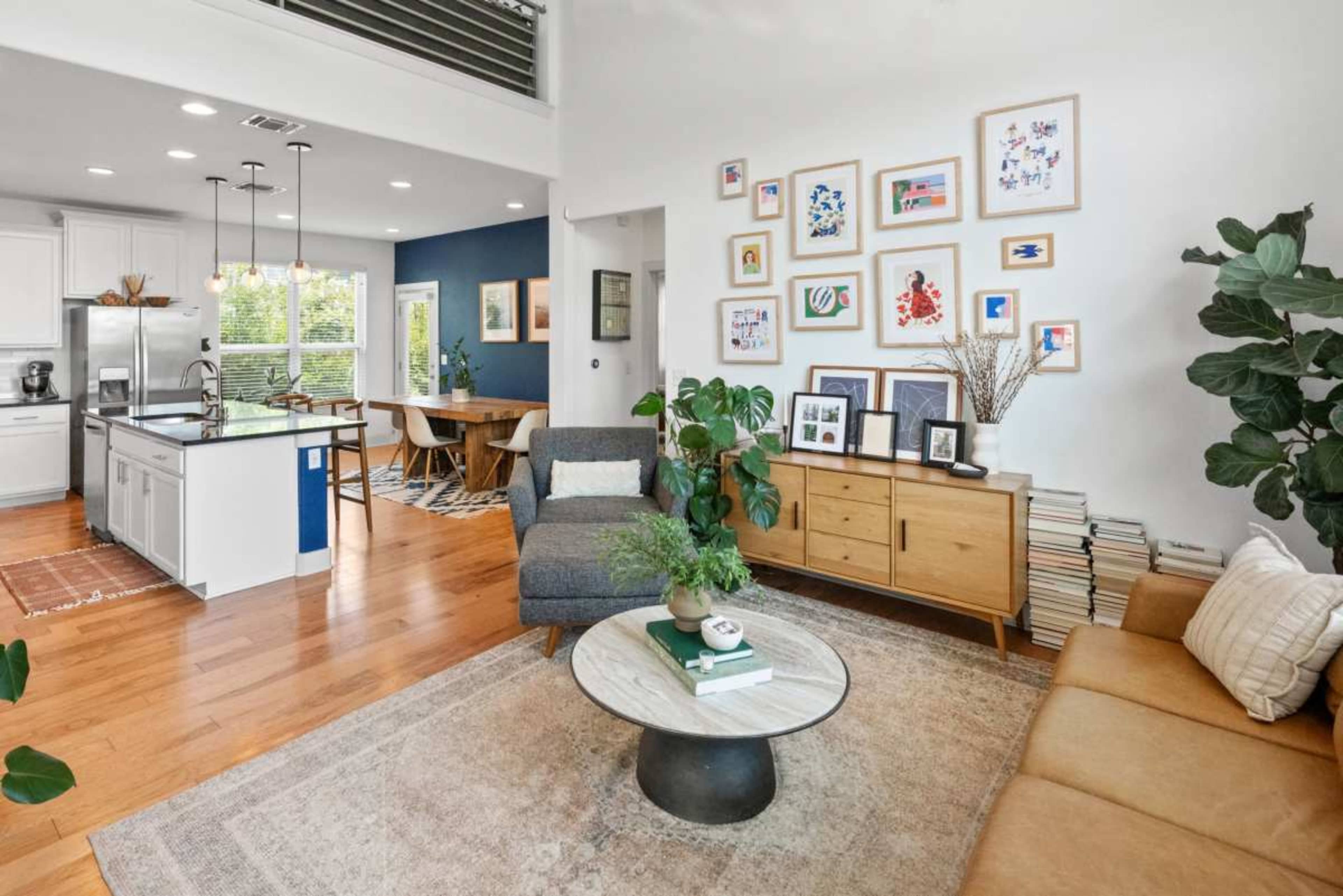 The image shows a modern living space featuring a seating area with a gray sofa and a tan couch, a round coffee table, and a dining area in the background with a wooden table and chairs, all surrounded by plants and artwork.
