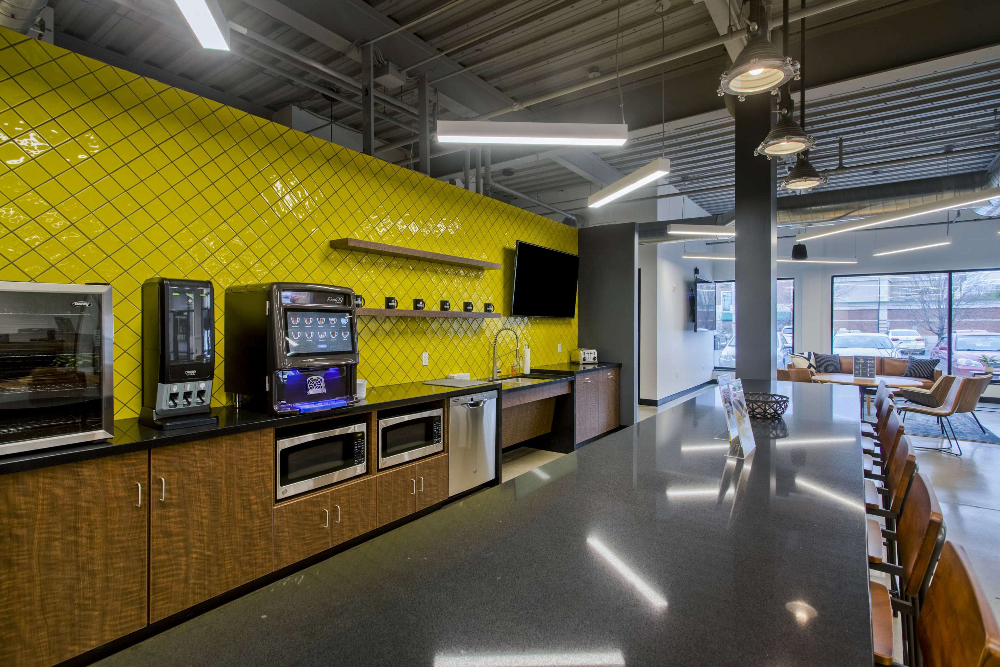 A modern kitchen area featuring yellow tiled walls, a counter with a coffee machine, vending machine, and two ovens, along with seating and large windows.