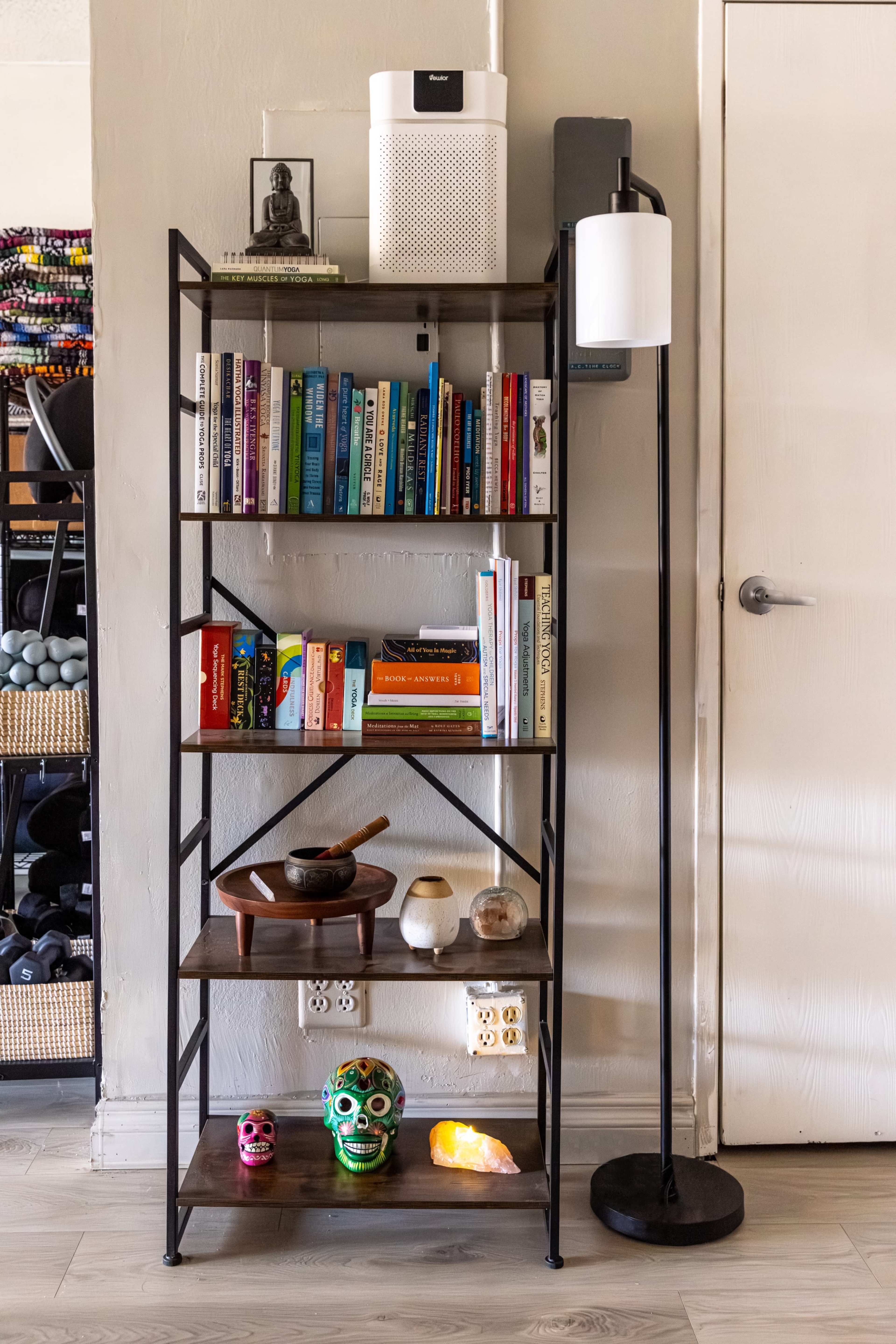A metal bookshelf with several rows of books, decorative items, and a lamp stands against a wall in a room.
