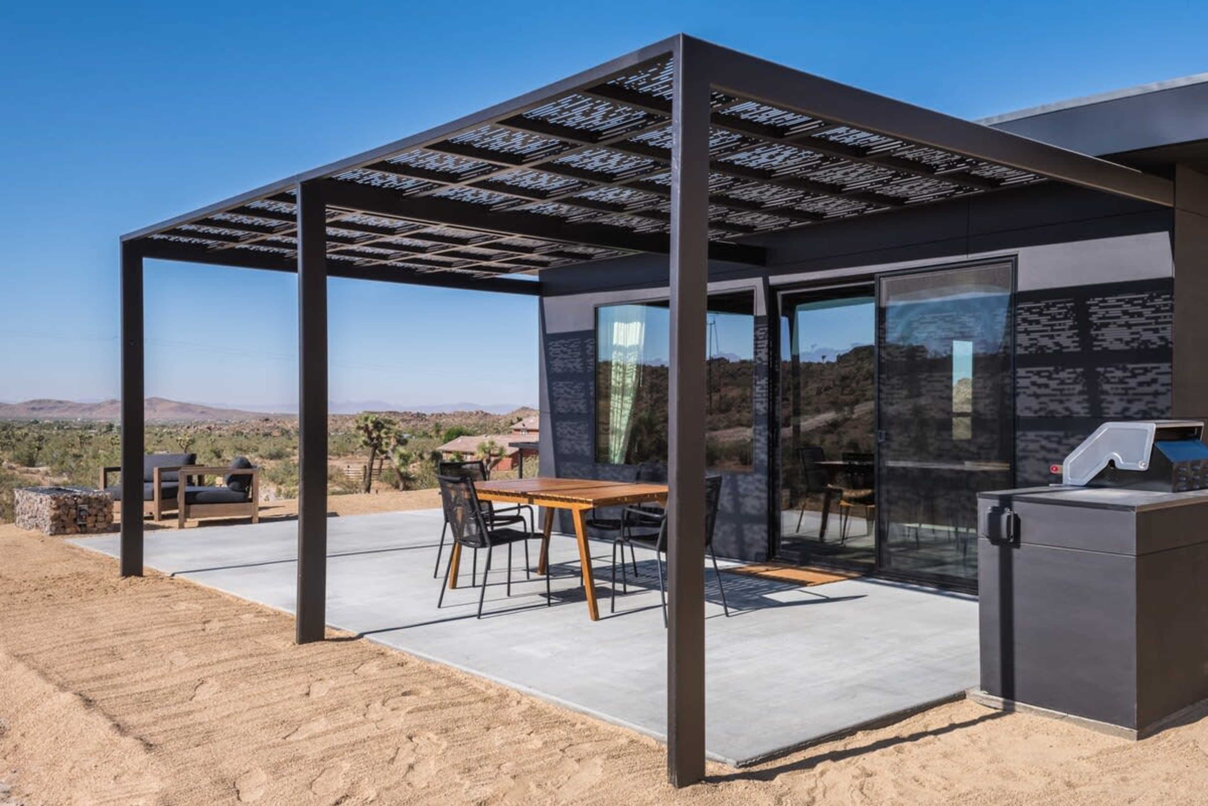 A modern black structure with a covered patio sits in a desert landscape, featuring a table and chairs under the shade.