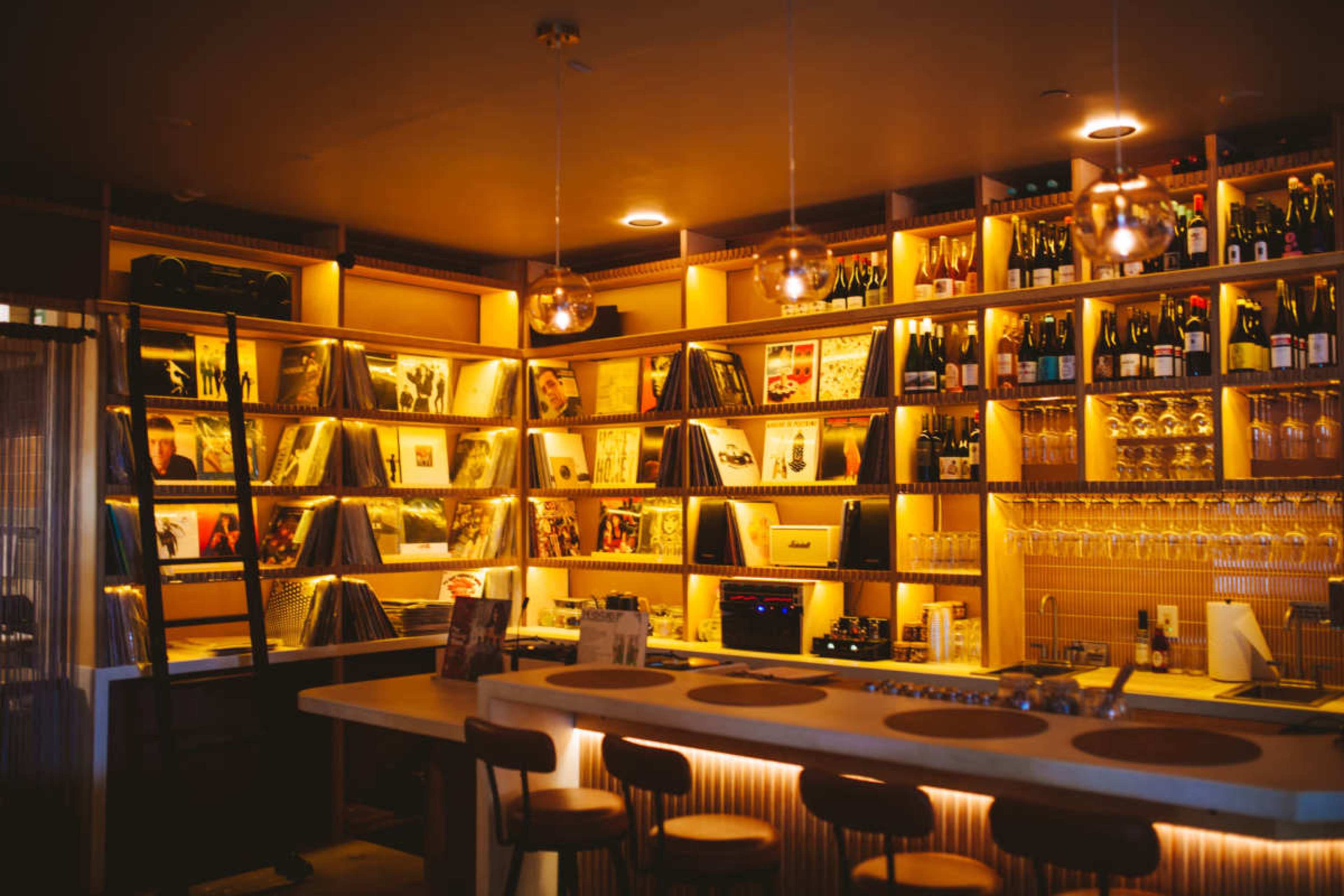 The image shows a dimly lit bar featuring shelves filled with vinyl records and various bottles of drinks, creating a cozy atmosphere.