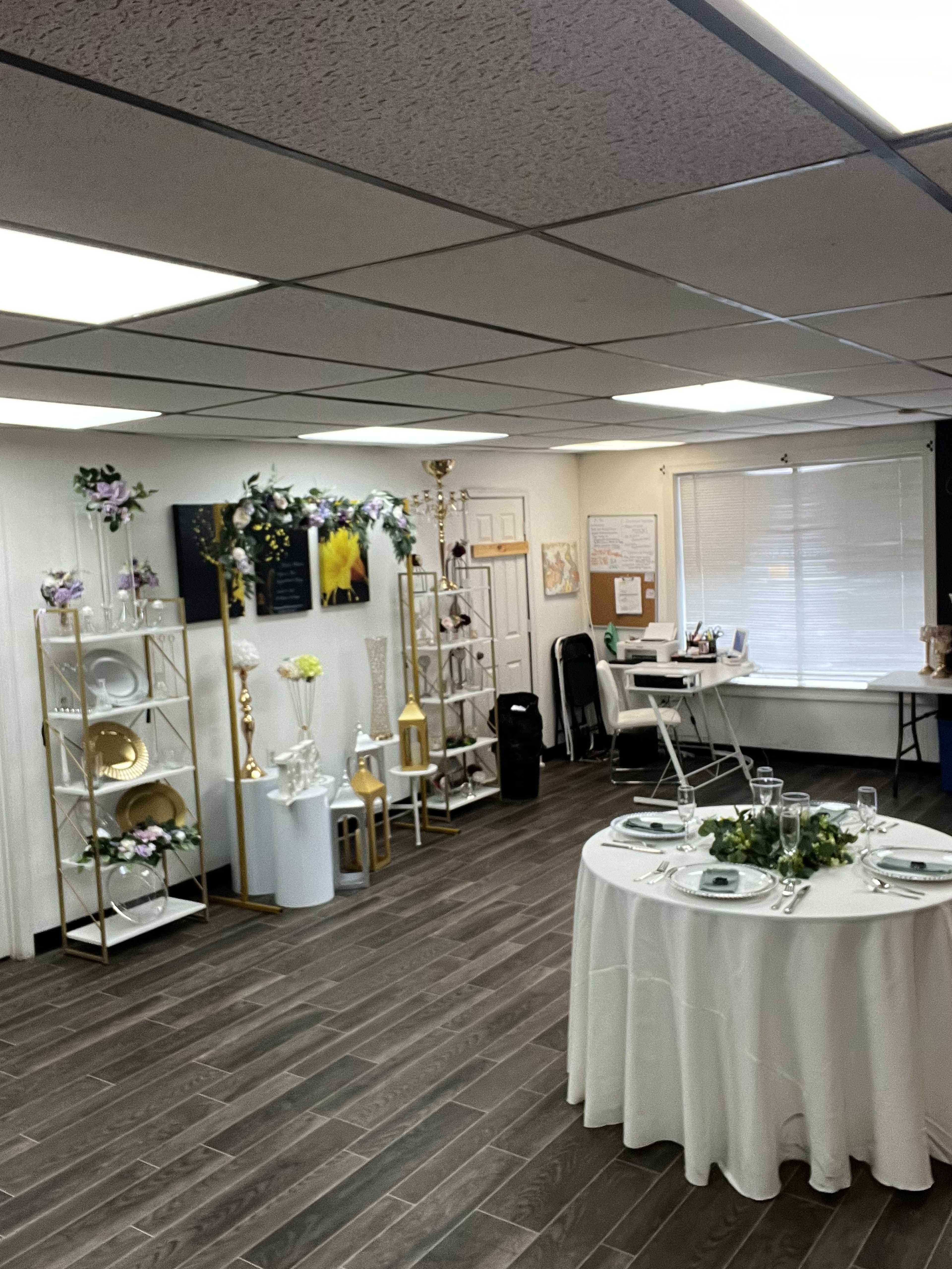 The image shows a decorated event space with a table set for a gathering, shelves displaying tableware, and various floral arrangements.